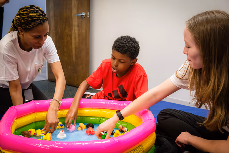 Two therapists and a small child playing at an inflatable pool, all seated in the center. The child is happily splashing water while the therapists assist and encourage them in their play. 