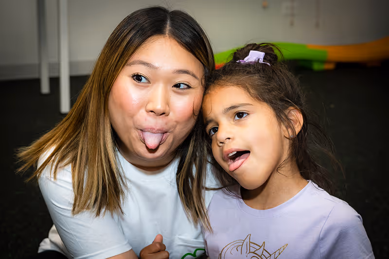 A behavior therapist and small child, smiling and sticking their tongues out. 