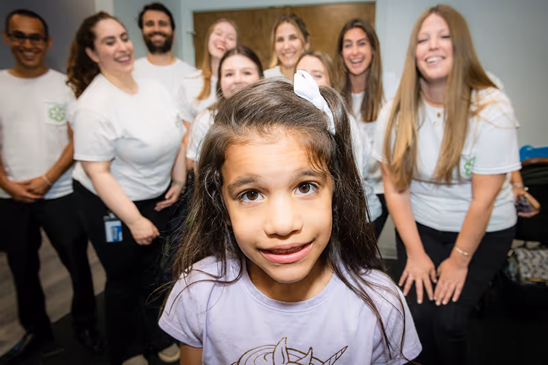 A smiling child with a group of pediatric therapists supporting in the background.