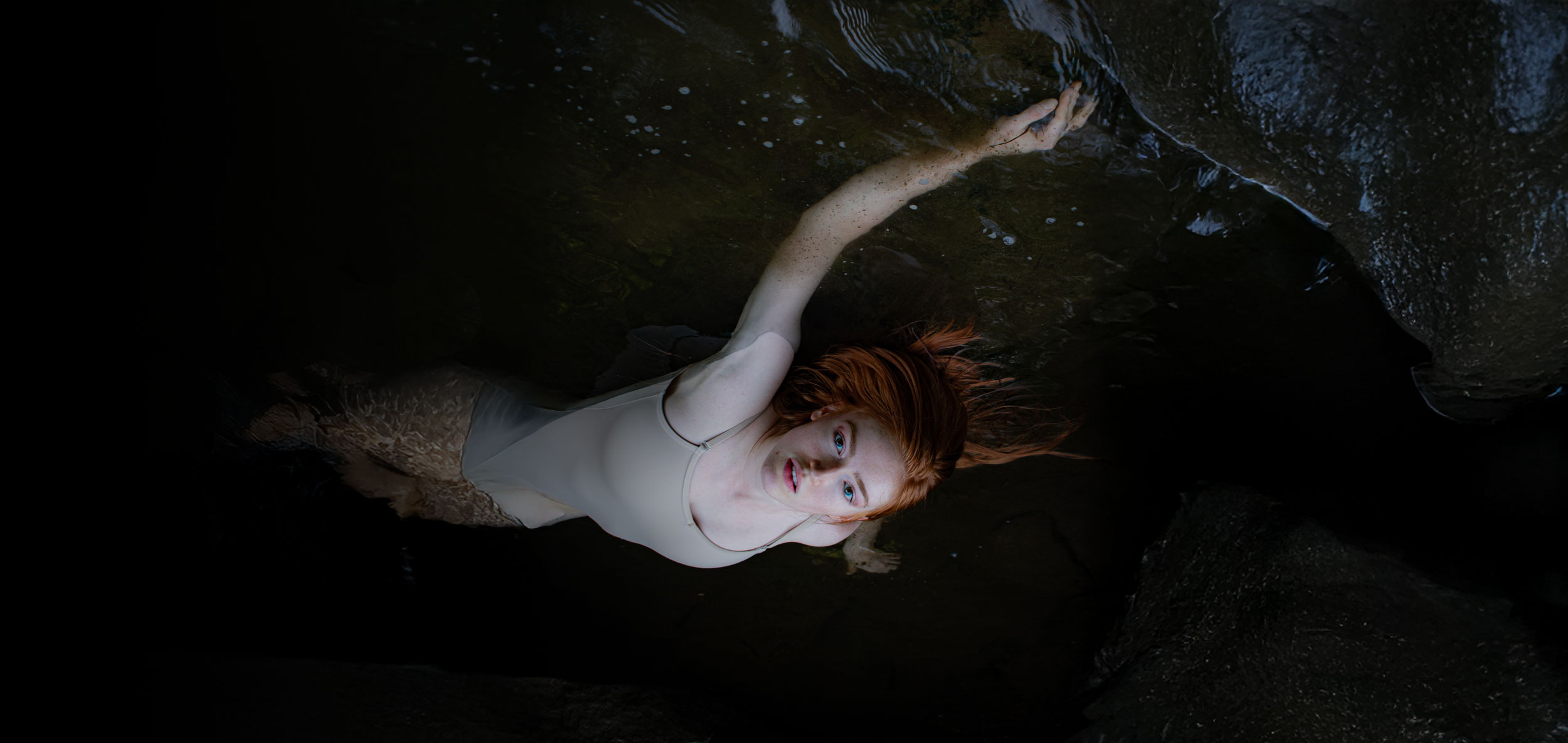 Red-haired woman in a beige swimsuit floating gracefully in dark water surrounded by rocks, with soft natural light highlighting her face and hair.