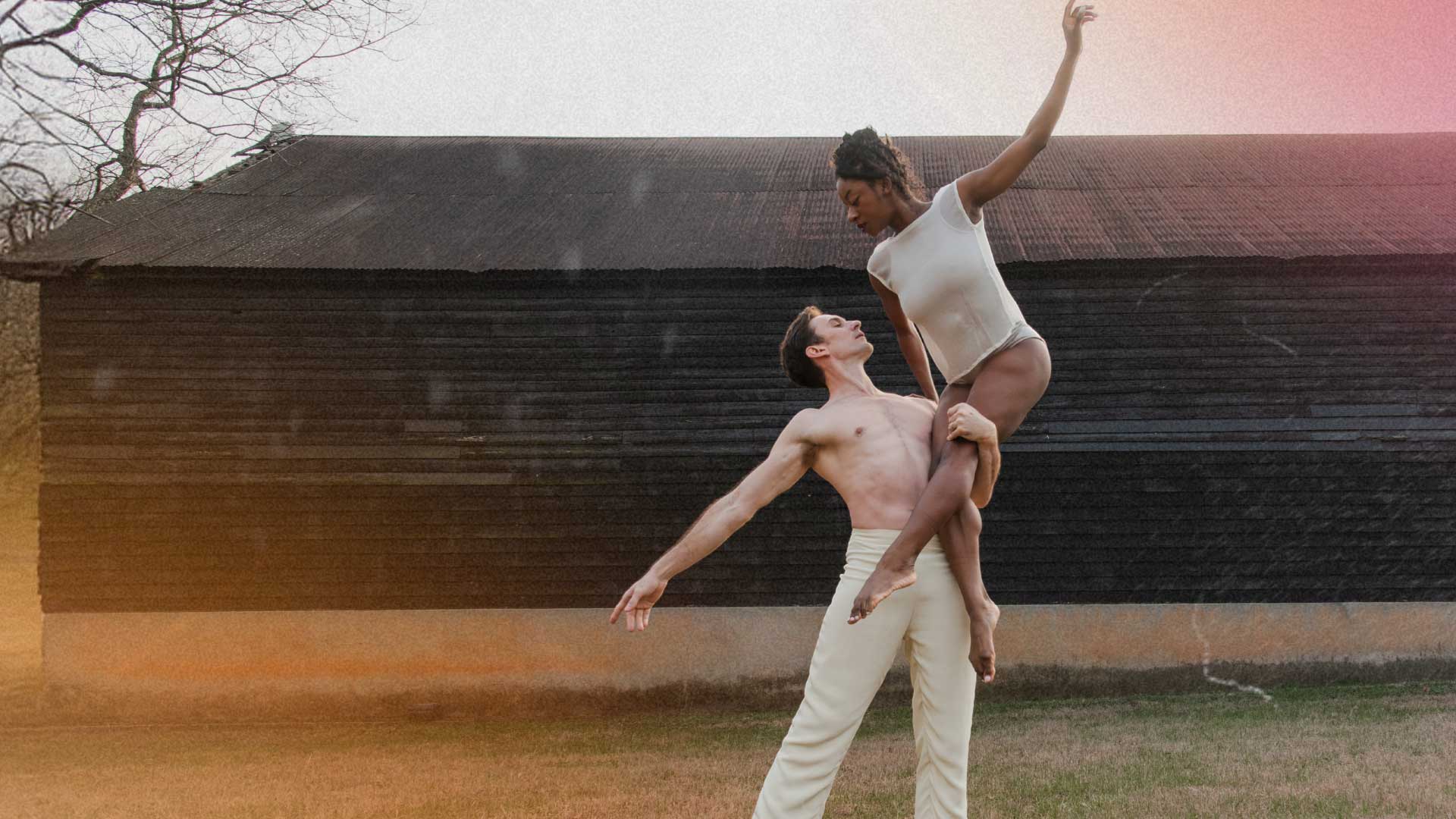 Two dancers perform an expressive lift outdoors in front of a rustic barn