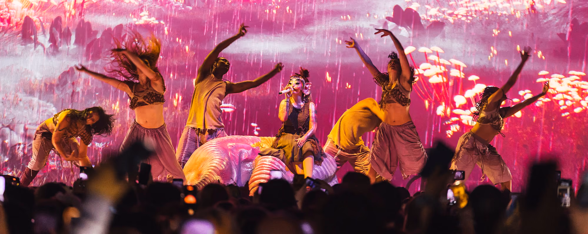Melanie Martinez in costume surrounded by dacners in front of a colorful pink other-worldy background