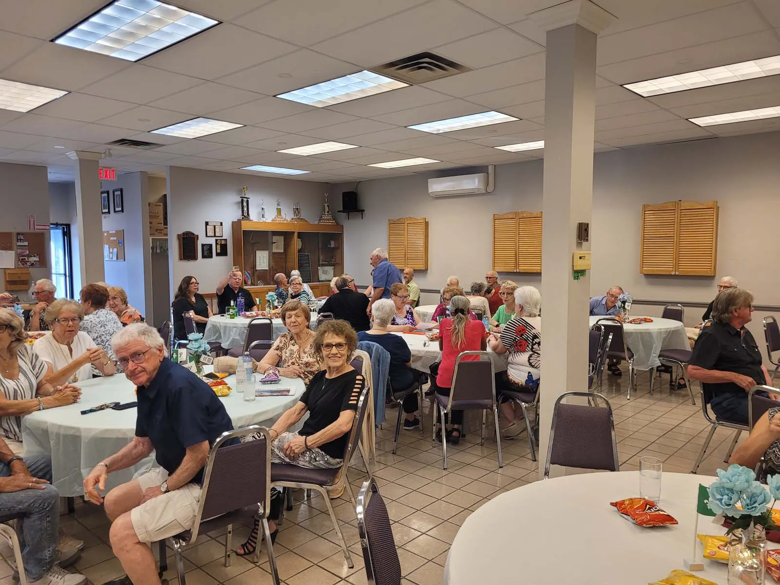 Vue d’ensemble de la salle communautaire remplie de participants assis aux tables rondes, profitant d’un moment convivial.