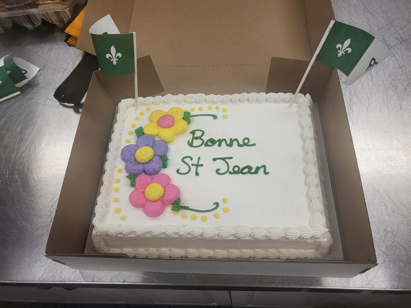 Un gâteau décoré avec des fleurs en crème et l’inscription « Bonne St Jean », accompagné de deux drapeaux verts avec la fleur de lys, prêt à être servi.