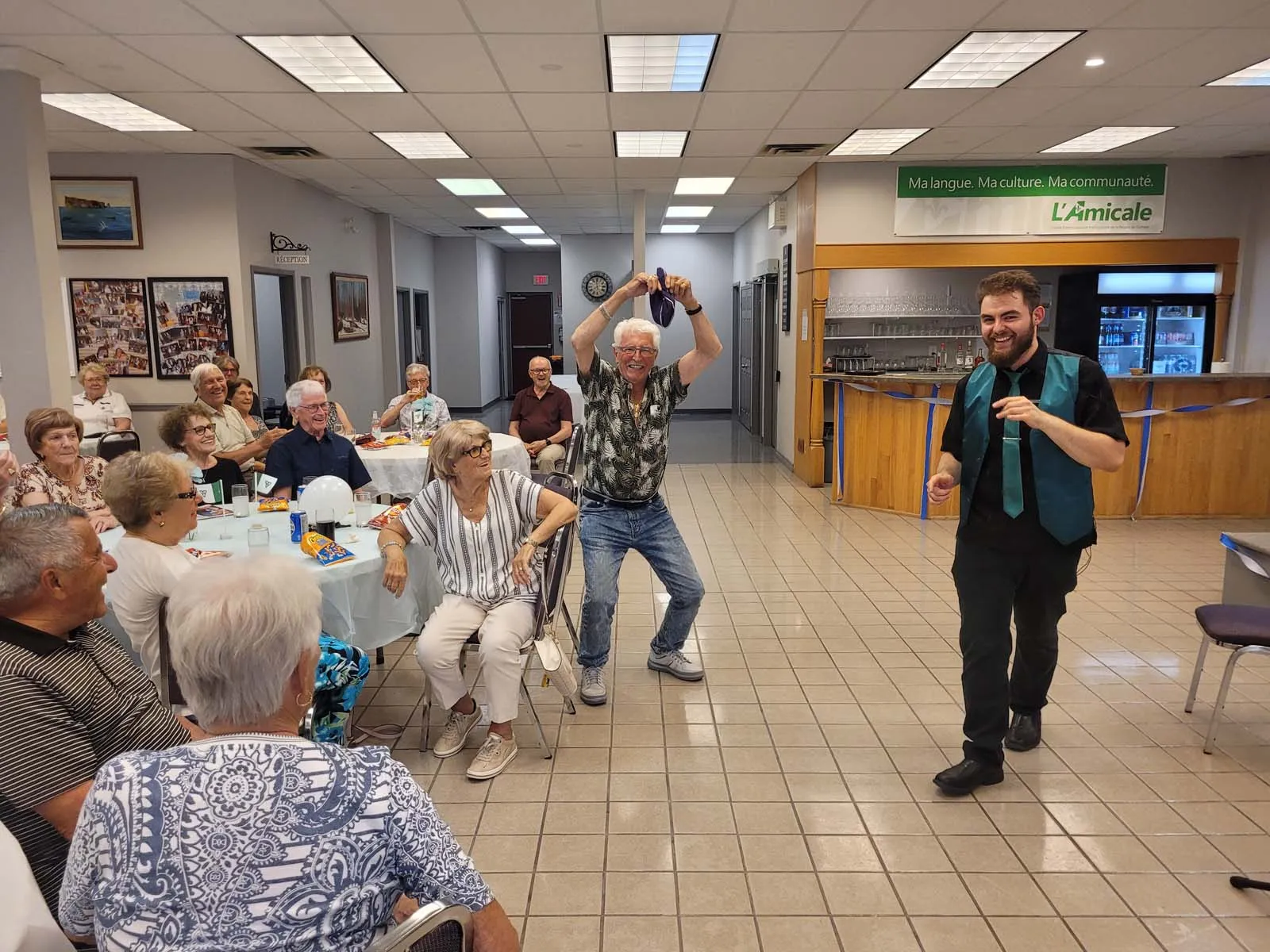 Un invité lève les bras en riant pendant que le magicien réagit, créant un moment joyeux sous les regards amusés de l’audience.