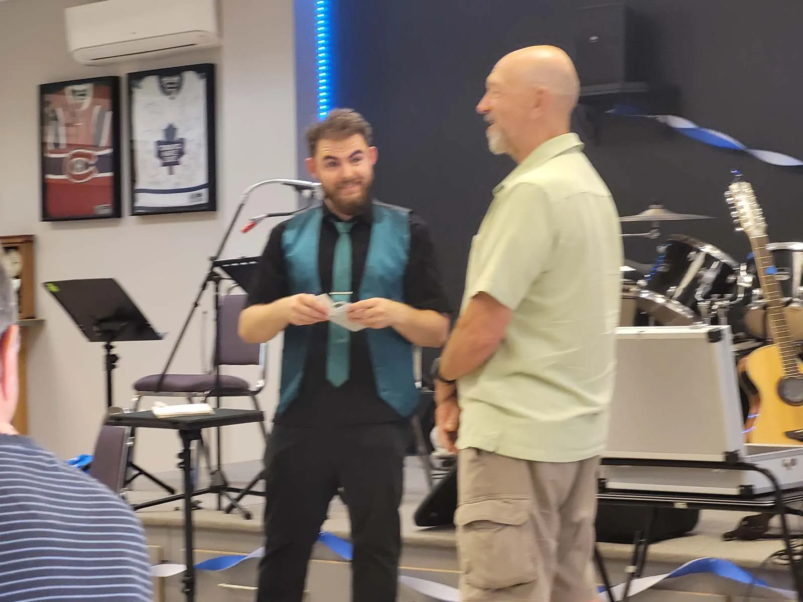 Le magicien effectue un tour sur scène avec un participant debout à ses côtés, tandis que les instruments en arrière-plan ajoutent à l’ambiance de spectacle.
