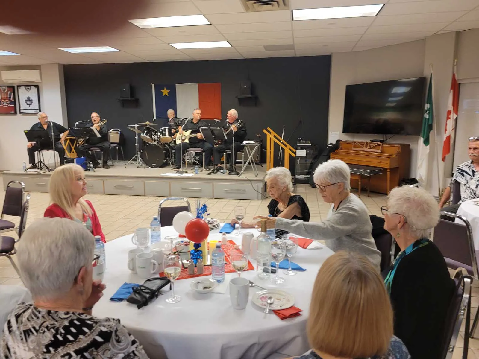 Les musiciens sur scène jouent pendant que les membres de L’Amicale partagent un repas à l’avant-plan. Les décorations bleu-blanc-rouge évoquent une célébration acadienne.Les musiciens sur scène jouent pendant que les membres de L’Amicale partagent un repa