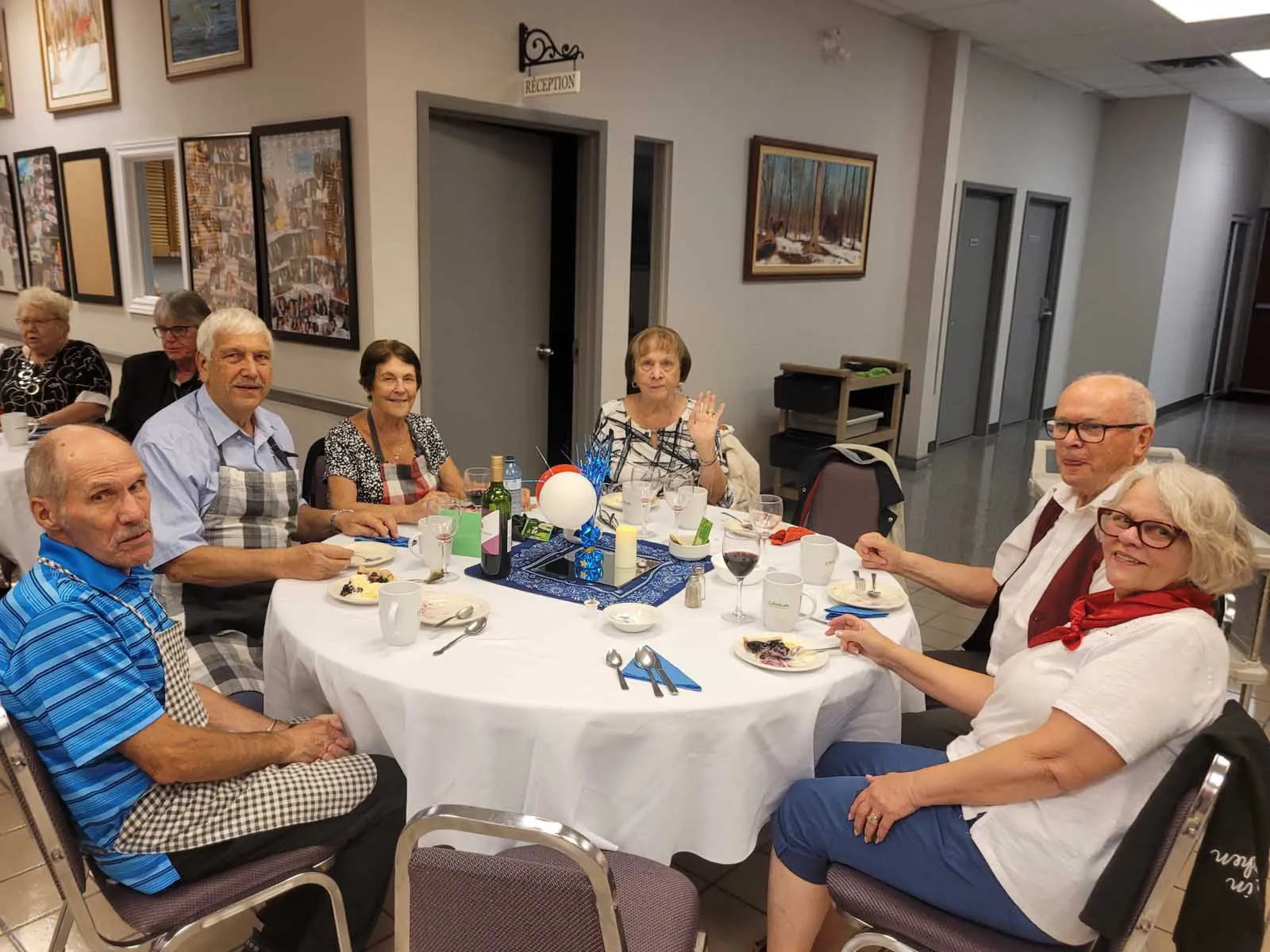Trois membres de L’Amicale sourient à la caméra à une table décorée de ballons et de serviettes rouges et bleues.