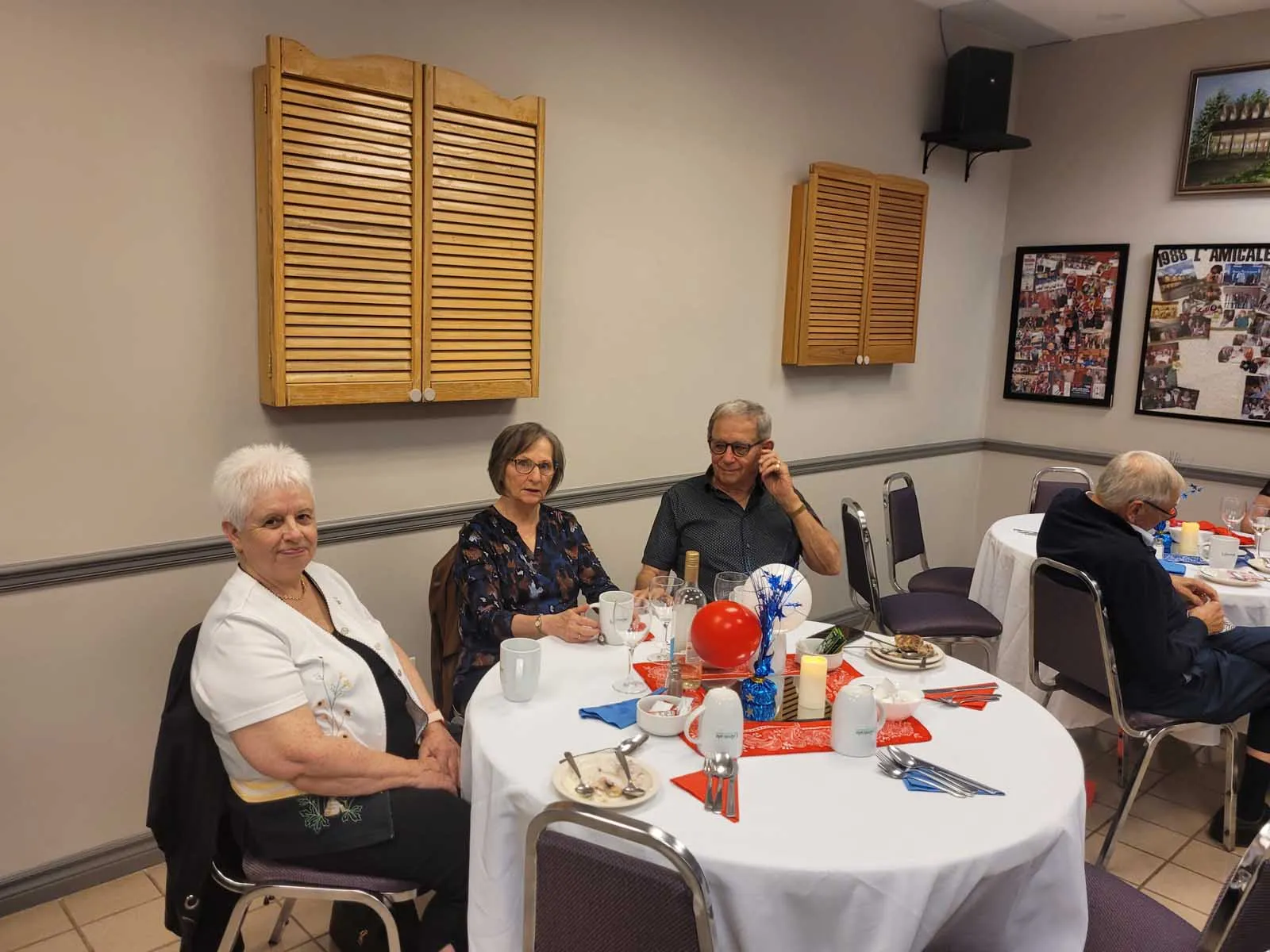 Des membres de L’Amicale assis à plusieurs tables rondes décorées de ballons rouges, en train de discuter et de profiter d’un repas convivial.