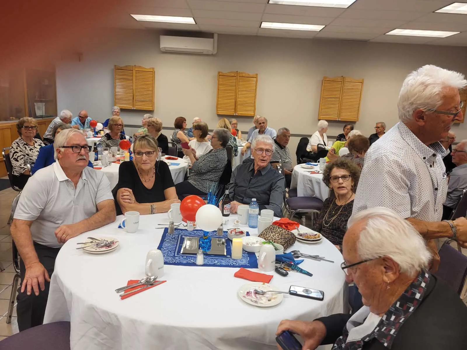 Une autre table de convives, cinq personnes souriantes posent devant un mur décoré de cadres photo retraçant la vie associative.