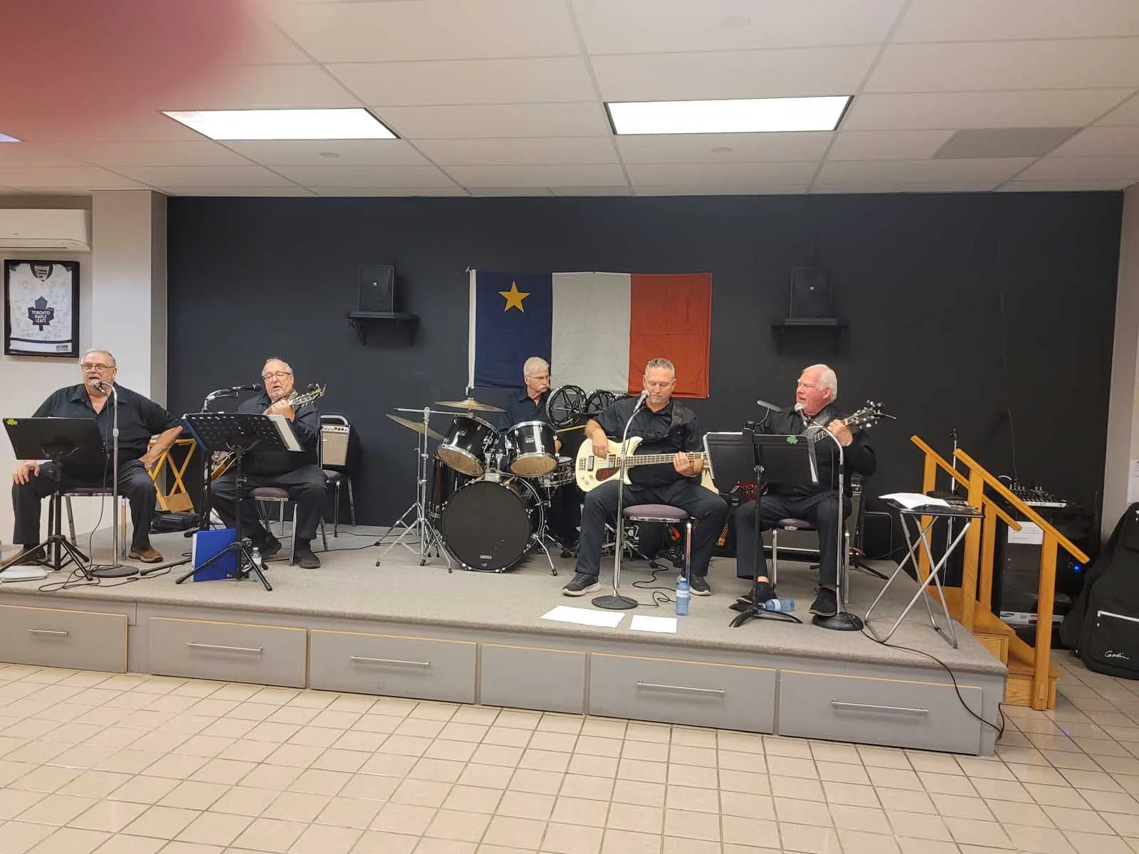 Un groupe de cinq musiciens, tous vêtus de noir, joue sur scène devant un drapeau acadien suspendu au mur.