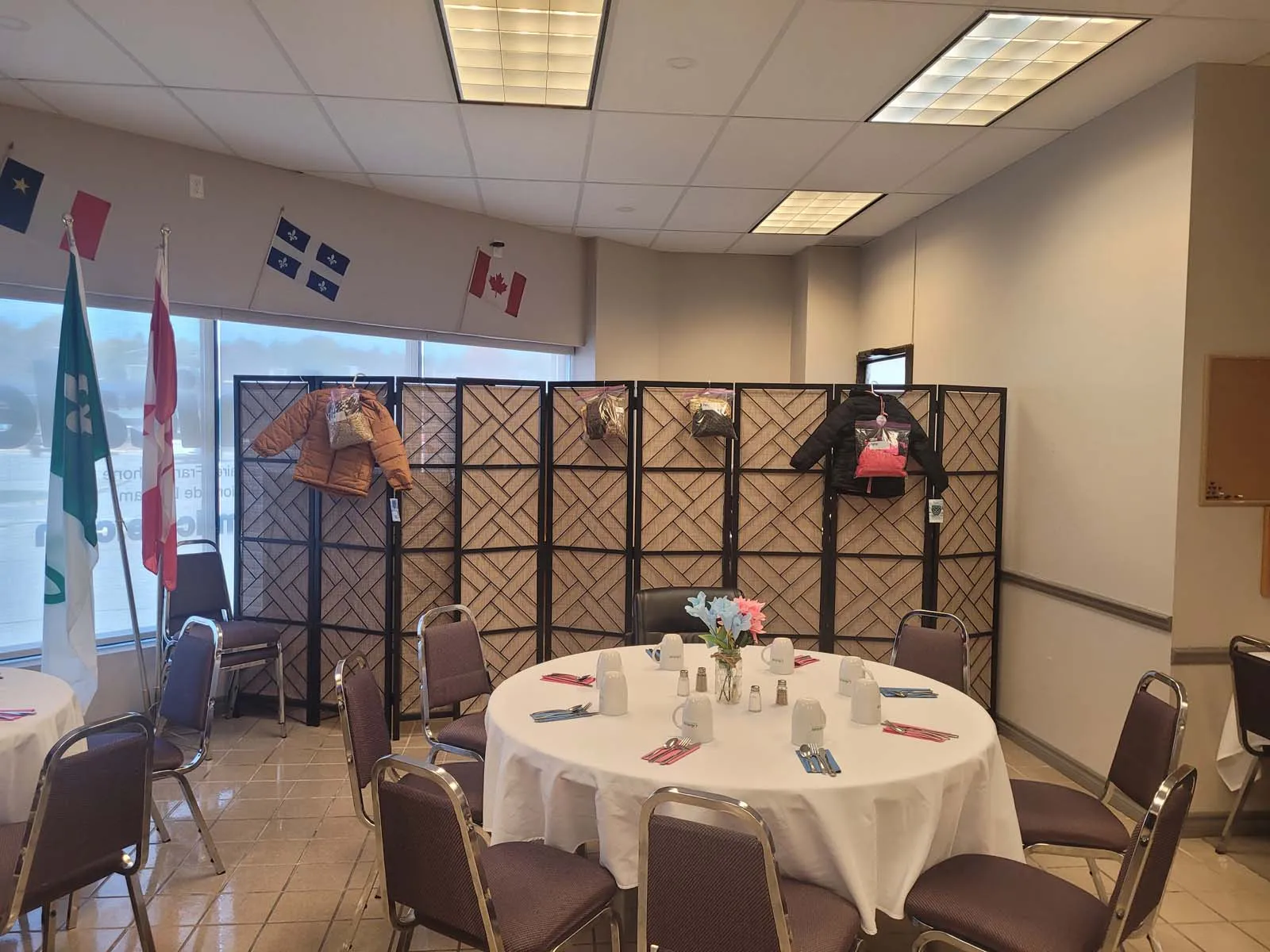 Salle communautaire décorée avec des drapeaux francophones et canadiens, montrant des manteaux pour enfants accrochés à un paravent derrière une table ronde joliment dressée, prête à accueillir les membres.