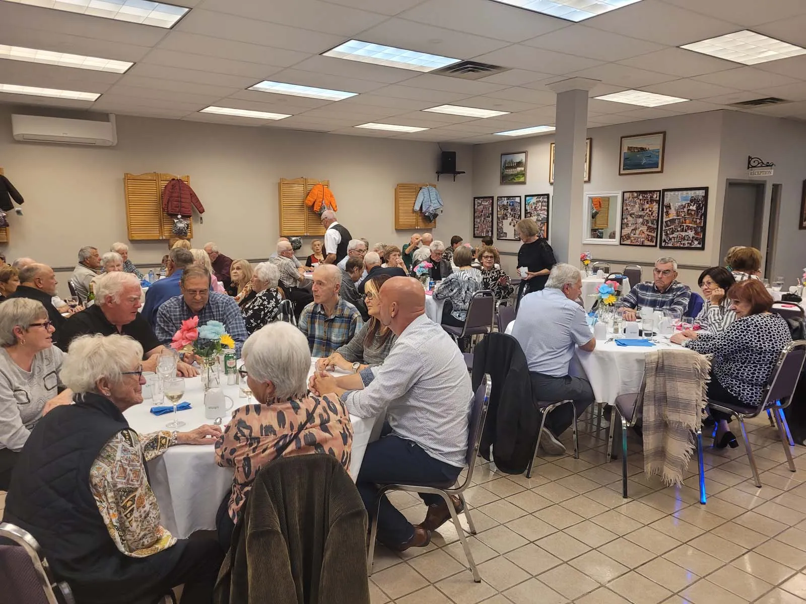 Membres de L’Amicale réunis autour des tables, en pleine discussion dans une ambiance conviviale, avec des manteaux suspendus en arrière-plan et des décorations murales.