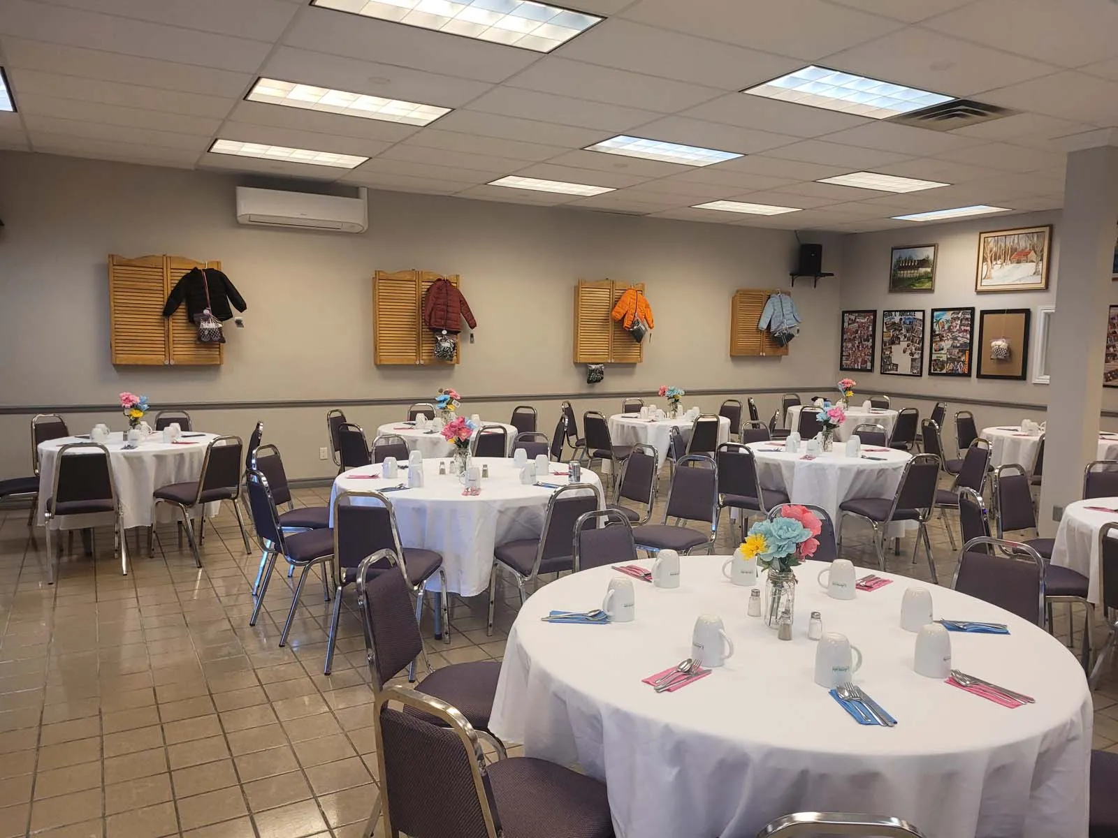 Vue d’ensemble de la salle préparée pour un événement de L’Amicale, avec plusieurs tables rondes dressées et des manteaux pour enfants exposés au mur, symbolisant une collecte ou une initiative communautaire.