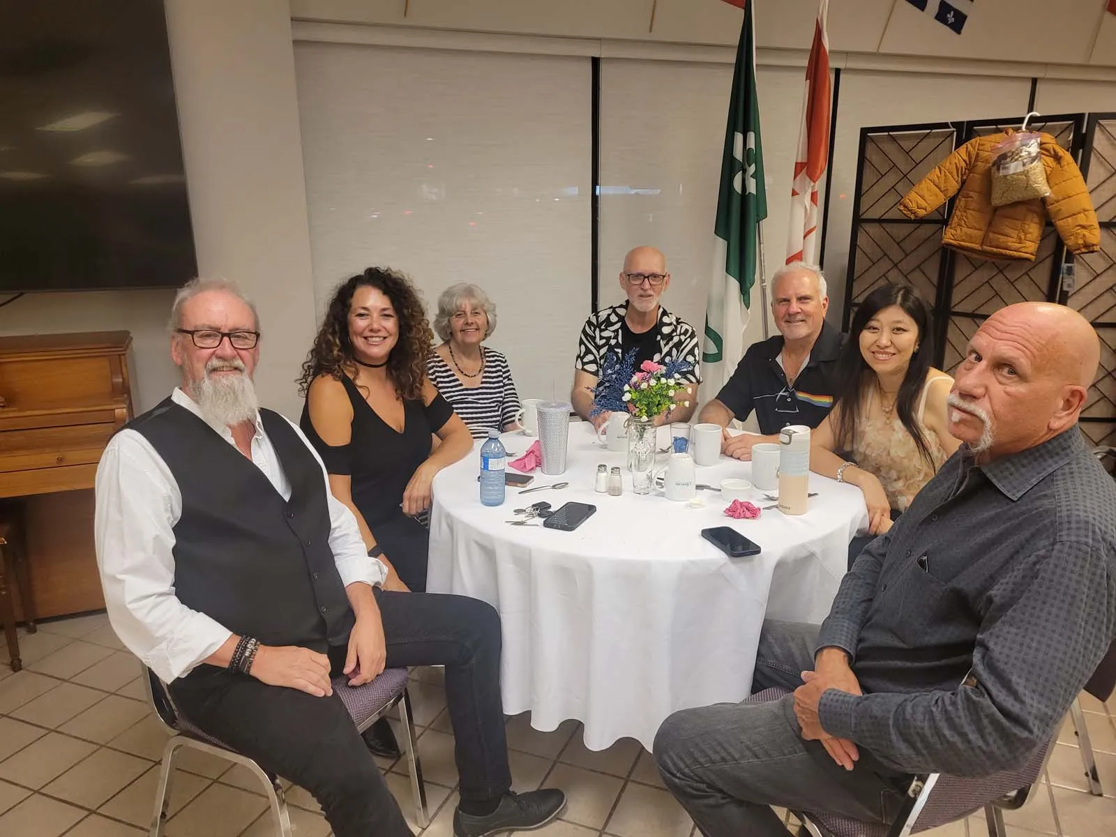 Groupe de membres de L’Amicale souriants, réunis autour d’une table décorée de fleurs, posant devant les drapeaux et le paravent portant un manteau pour enfant.