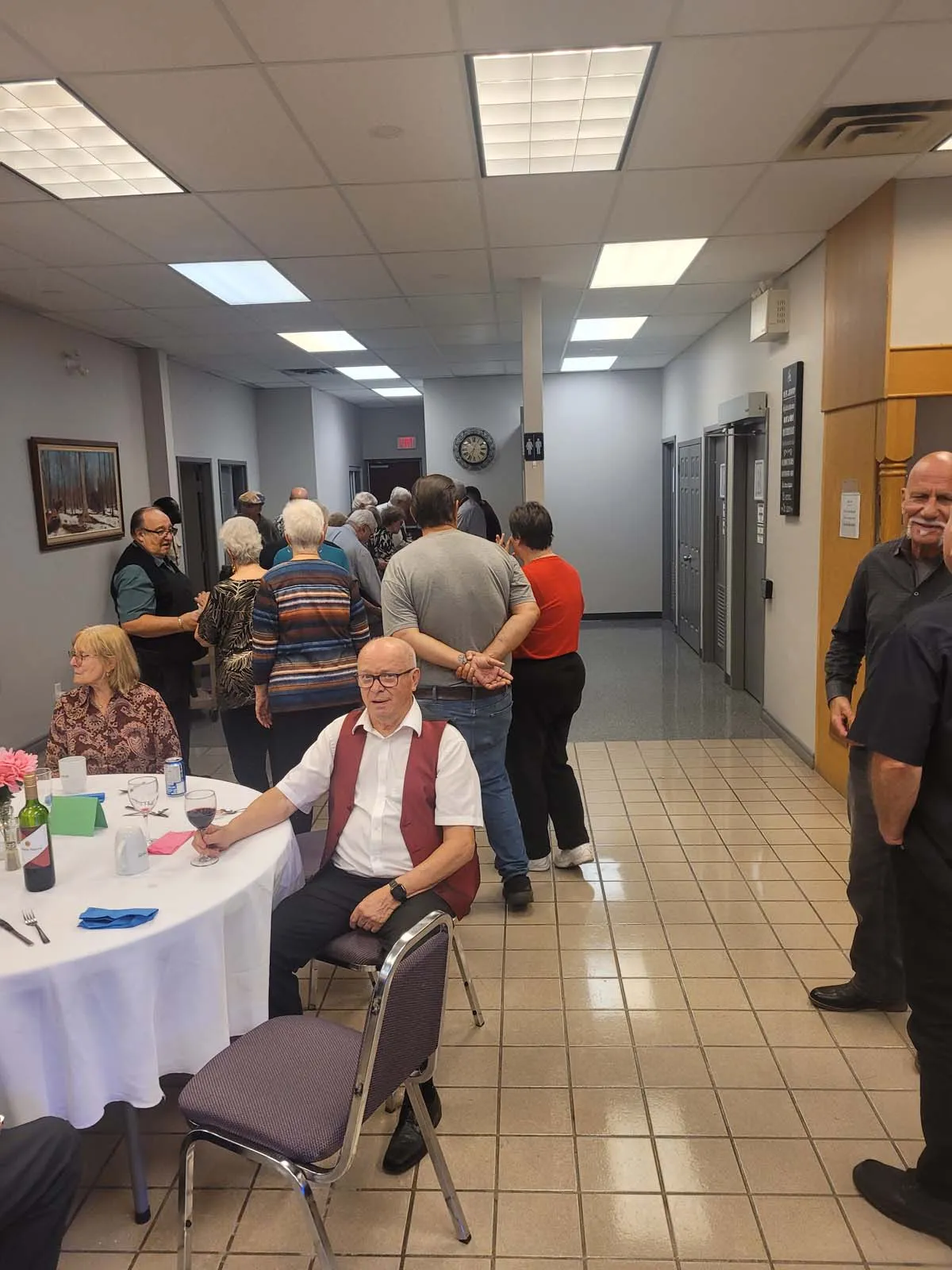 File d’attente de membres de L’Amicale dans le couloir menant au service de repas, pendant qu’un autre membre est assis avec un verre de vin à une table.