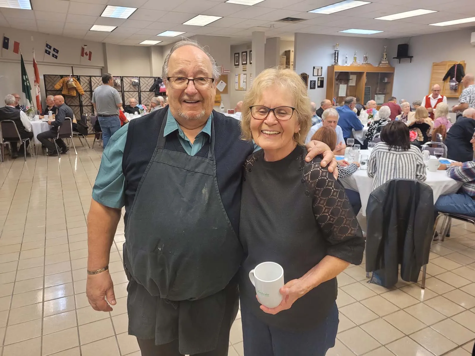Deux membres souriants de L’Amicale posent côte à côte dans la salle, entourés de participants assis aux tables décorées.