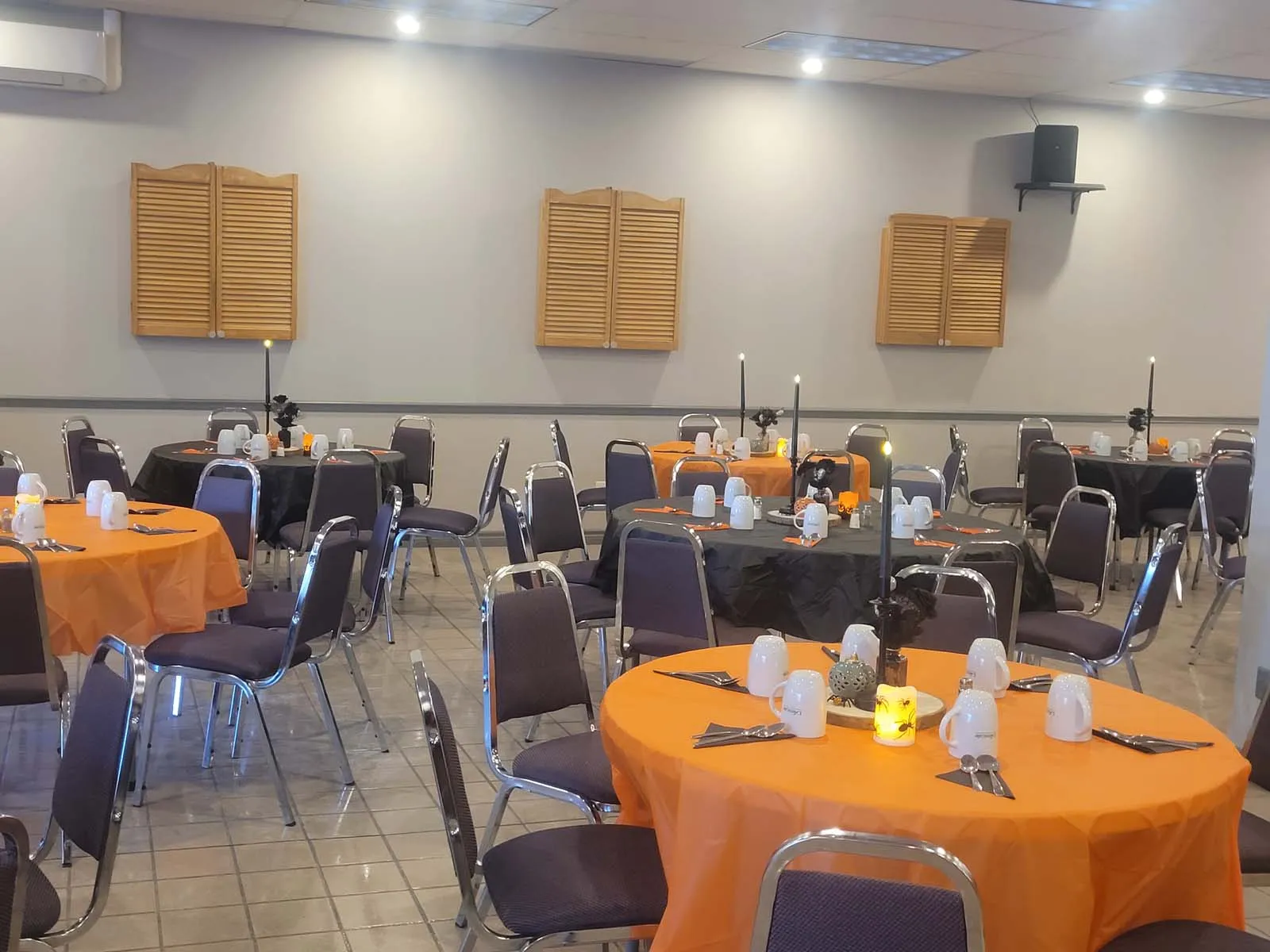 Vue de la salle de réception décorée pour l’Halloween avec des nappes orange et noires, des chandelles LED sur les tables, et des tasses blanches alignées avec soin.