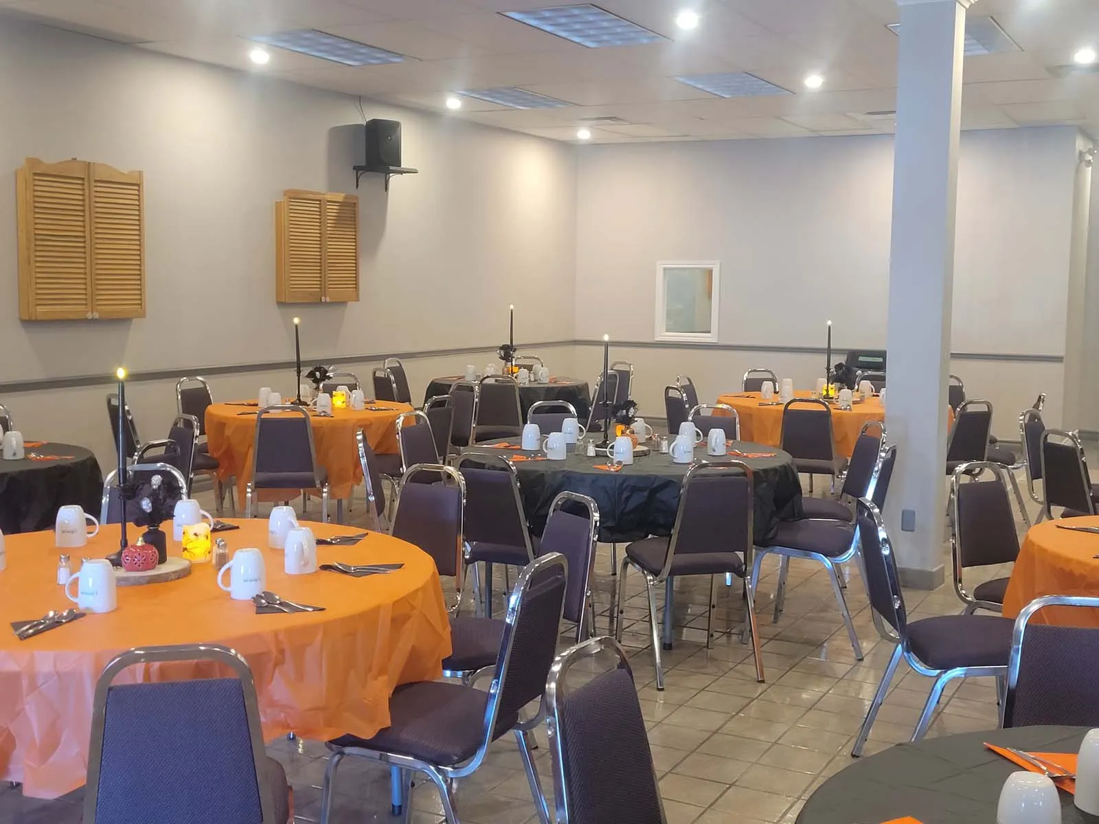 Disposition de plusieurs tables rondes recouvertes de nappes orange et noires, avec des décorations de citrouilles lumineuses et chandelles sombres au centre, prêtes à accueillir les membres pour un souper thématique.