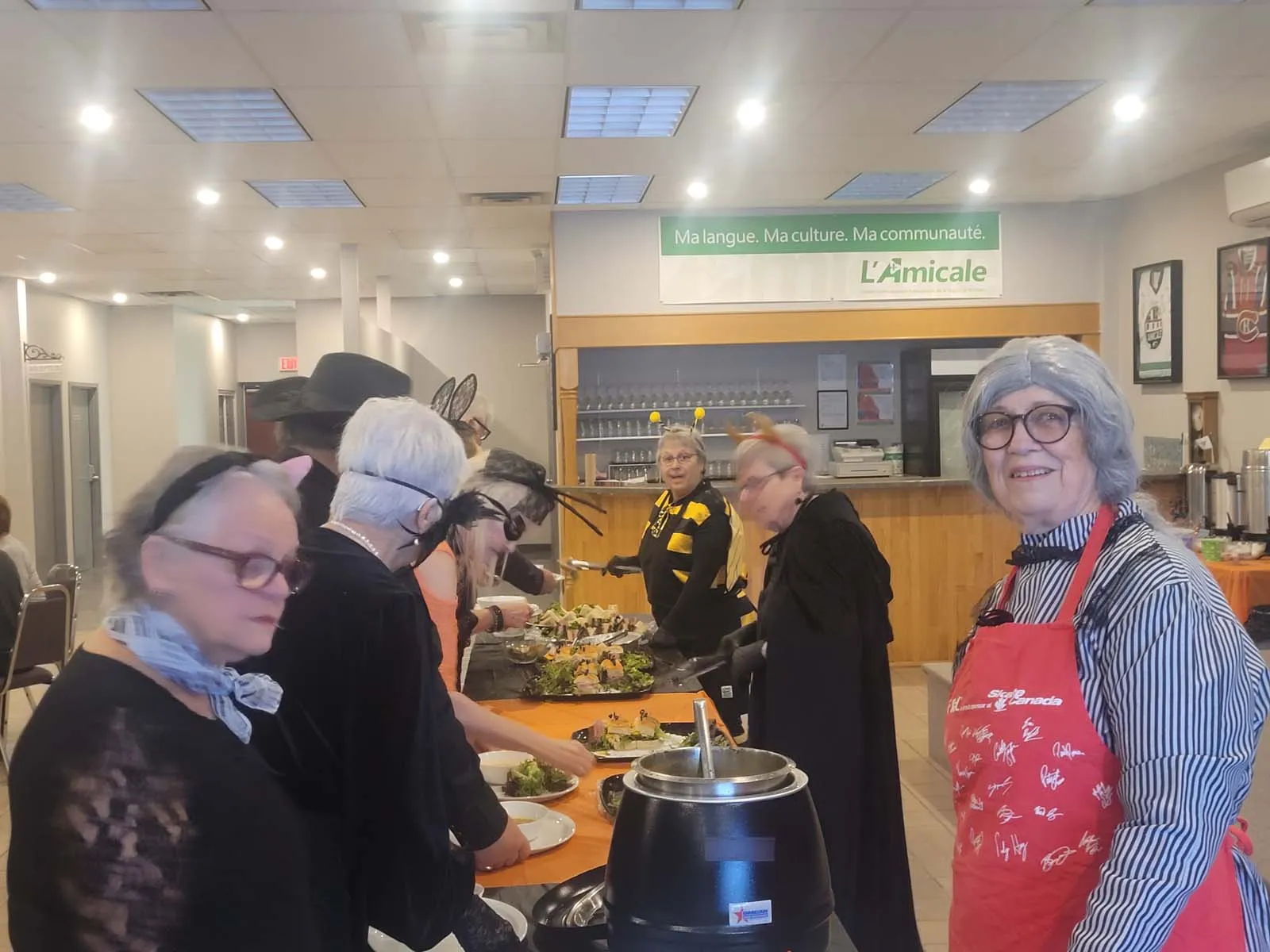 Groupe de membres costumés servant de la soupe et des plats variés à un buffet. L’ambiance est festive avec des déguisements de sorcières, abeilles et autres personnages.