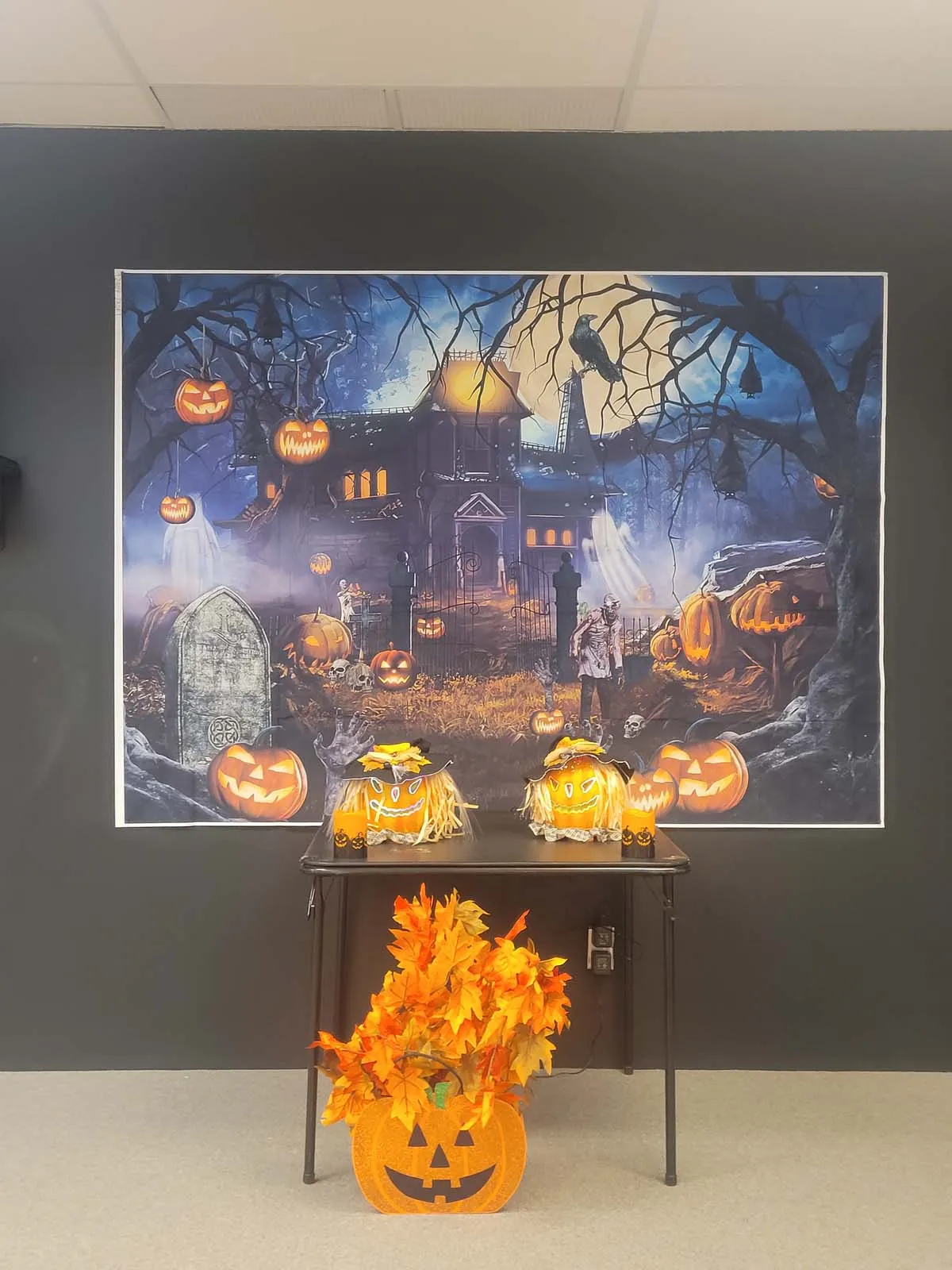 Un grand décor mural représentant un manoir hanté éclairé par une pleine lune, avec des citrouilles lumineuses et des arbres sinistres. Devant, une table noire ornée de citrouilles décoratives et d’un pot de feuilles d’automne orange en forme de citrouille