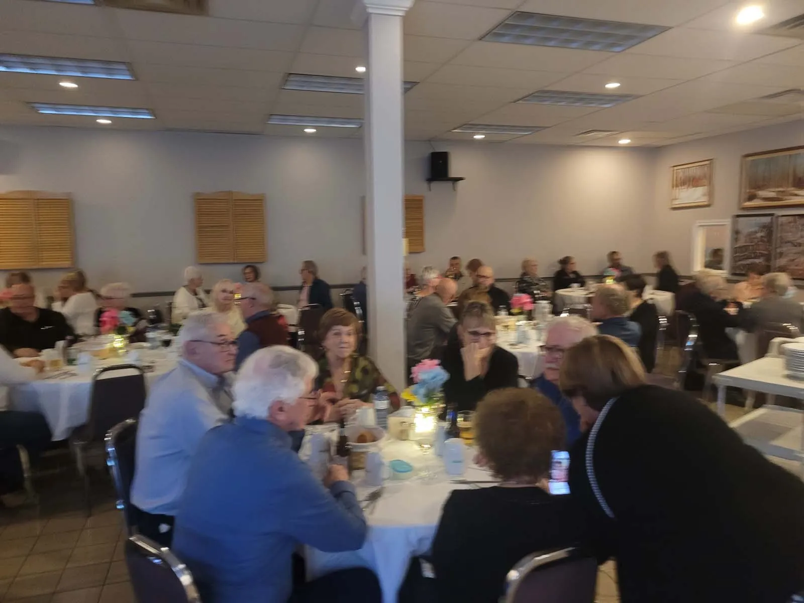 Vue floue et animée d’une salle remplie de convives installés à des tables rondes, ornées de fleurs et baignées d’un éclairage doux.