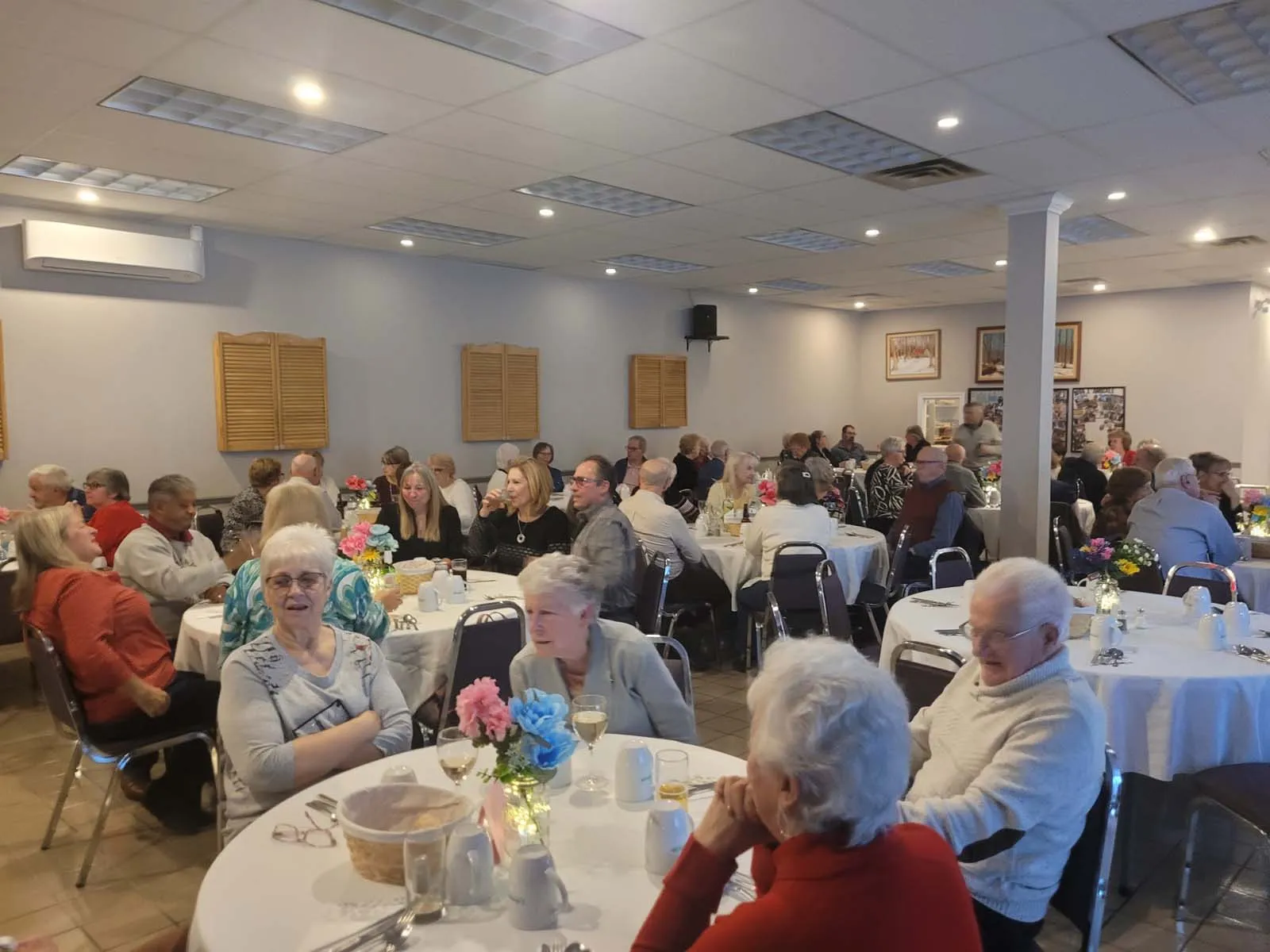 Un grand groupe de personnes âgées assises à des tables rondes, discutant et partageant un moment convivial dans une ambiance chaleureuse.