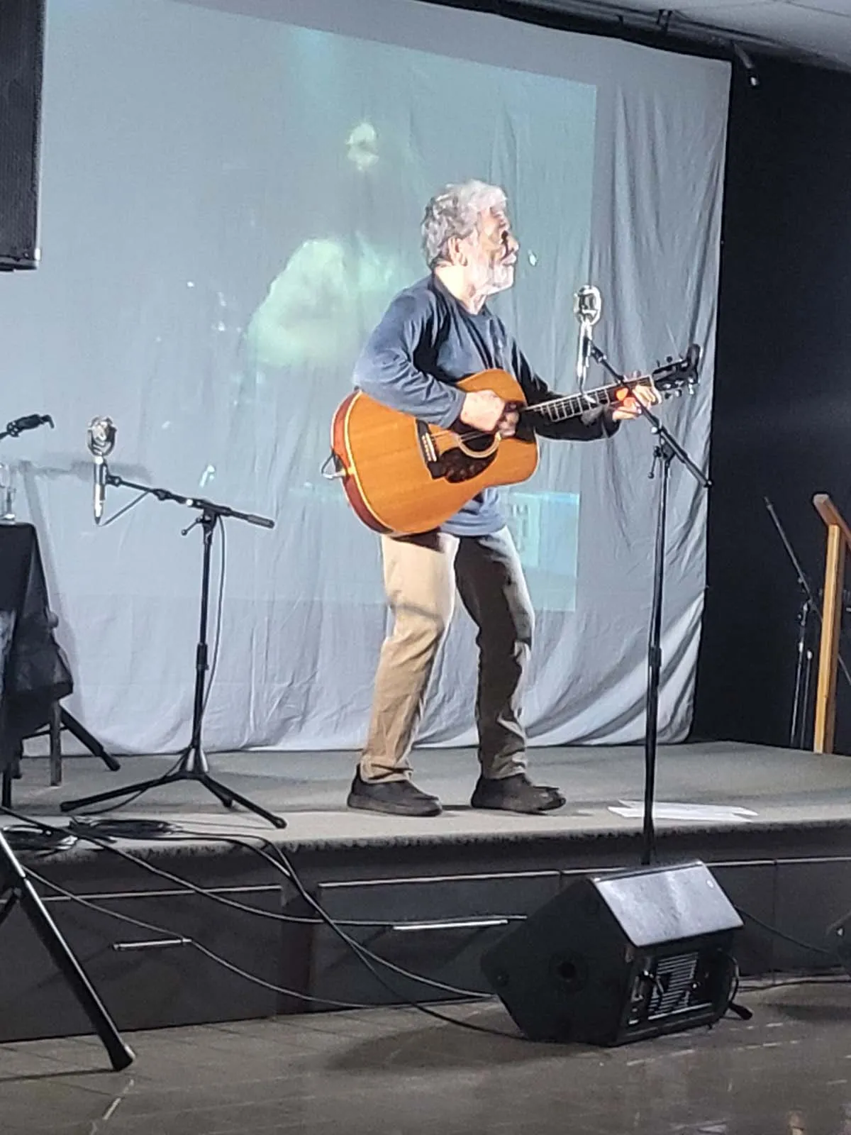 Un homme aux cheveux gris joue de la guitare acoustique sur scène devant un écran de projection affichant son image.