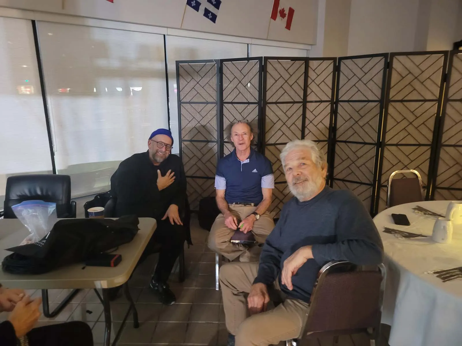 Trois hommes souriants assis devant un panneau pliant, dans une salle décorée de drapeaux suspendus.