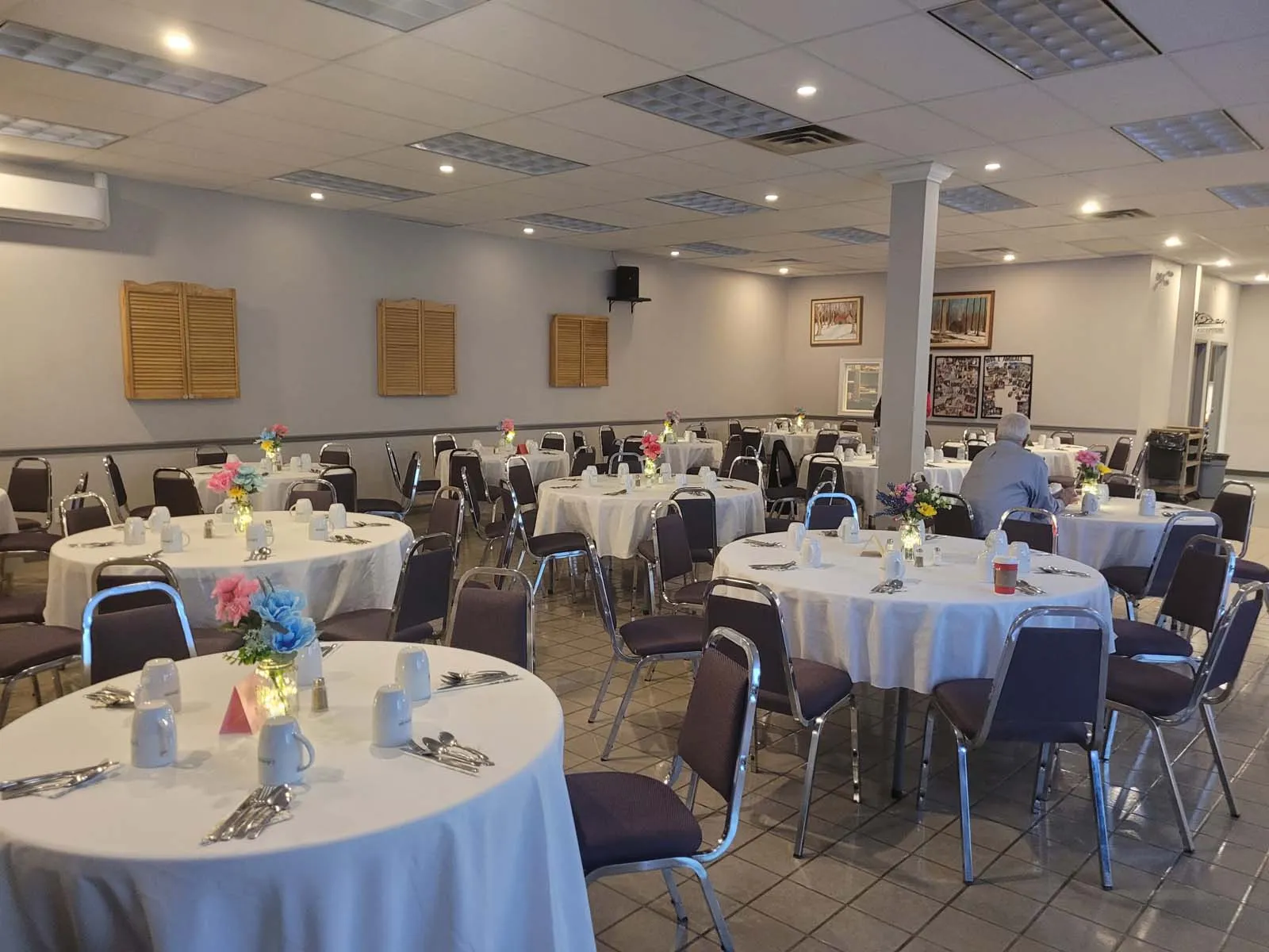 Vue d’une salle de banquet avec plusieurs tables rondes couvertes de nappes blanches, décorées de centres de table floraux colorés et de vaisselle prête pour un repas.