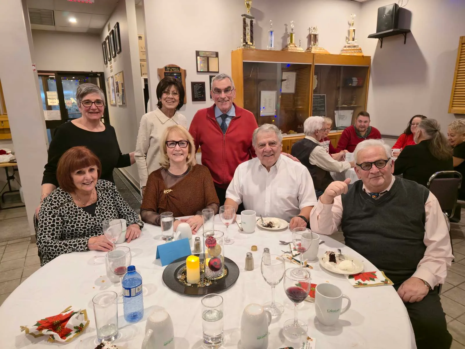 Groupe de membres de L’Amicale posant ensemble près d’une table, avec une vitrine de trophées en arrière-plan, dans une ambiance chaleureuse.