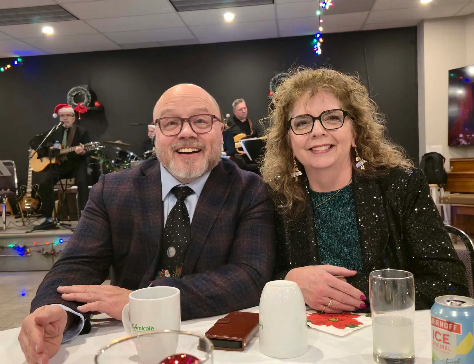 Deux membres de L’Amicale souriant à la caméra, assis à une table devant la scène où un groupe de musique joue en arrière-plan.
