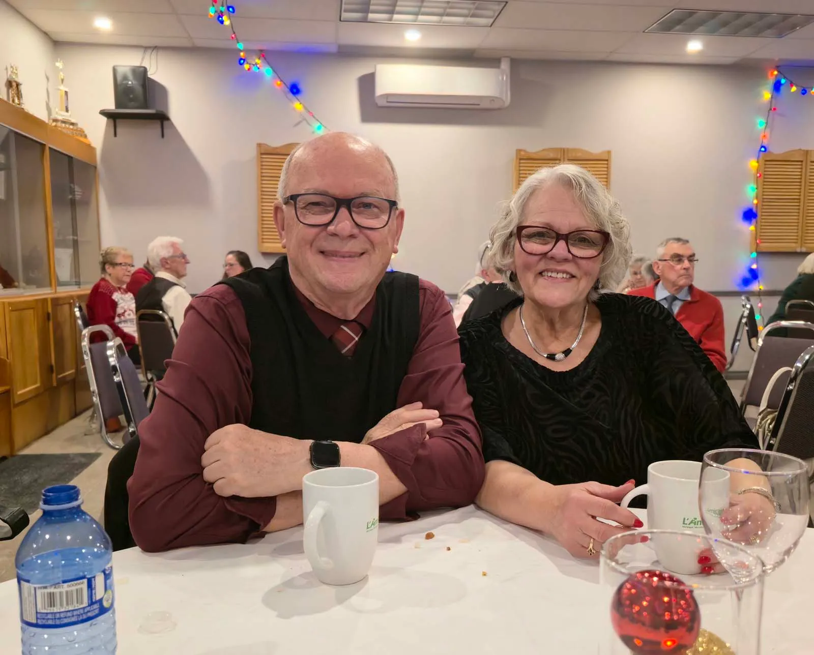 Deux membres de L’Amicale attablés ensemble, souriant à la caméra, avec des tasses et décorations festives visibles sur la table.
