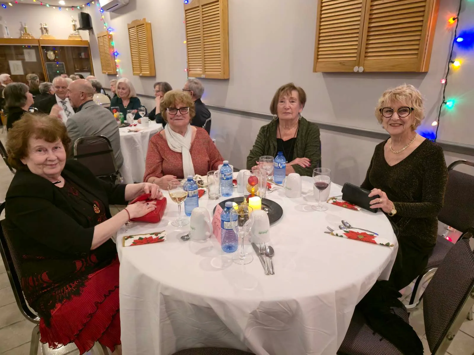 Quatre membres de L’Amicale assis à une table, souriant à la caméra avec des boissons et décorations festives au centre.