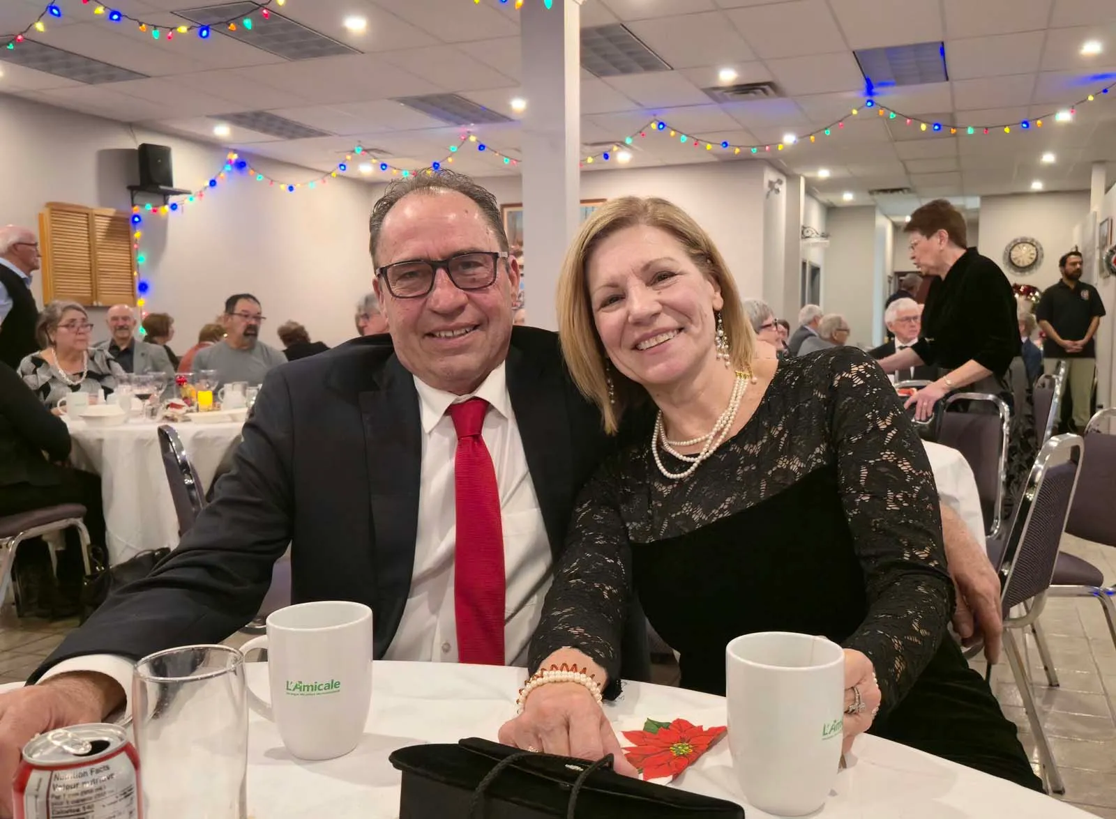Deux membres de L’Amicale assis côte à côte, souriant chaleureusement à la caméra dans la salle décorée pour l’événement.