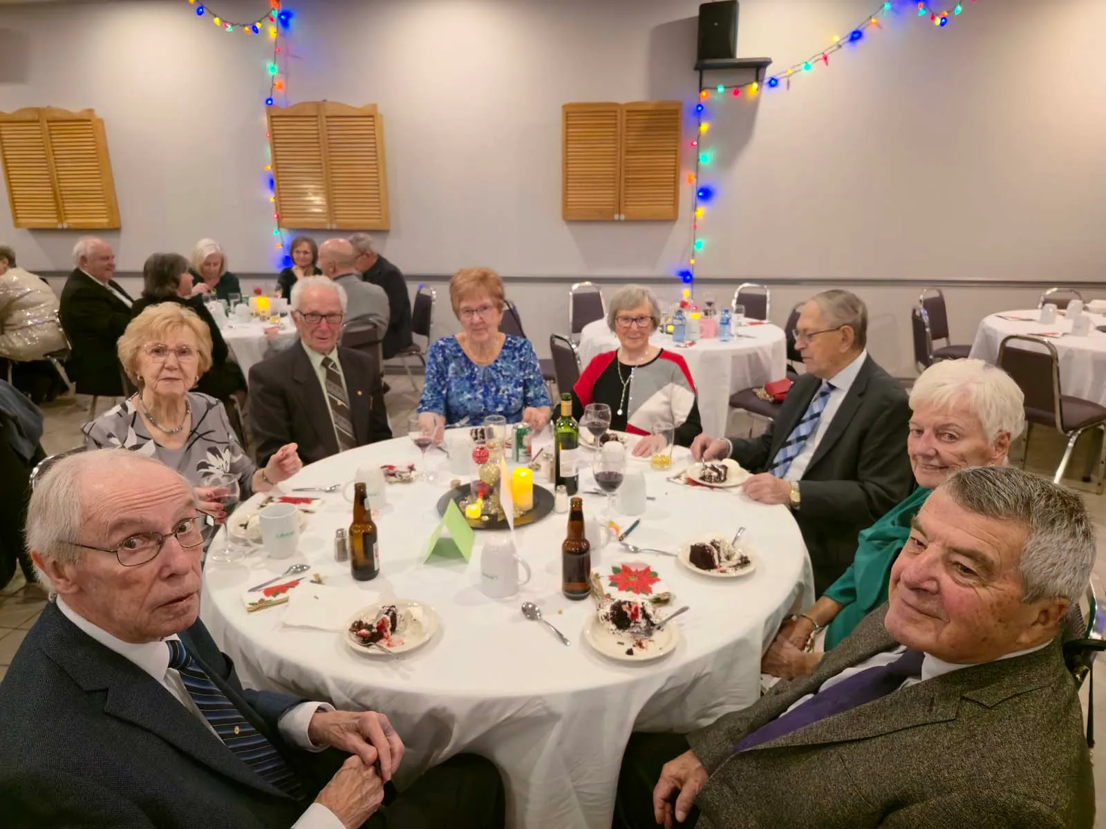 Table ronde avec huit membres souriants dégustant leur part de gâteau, dans une salle décorée de lumières de Noël suspendues au mur.