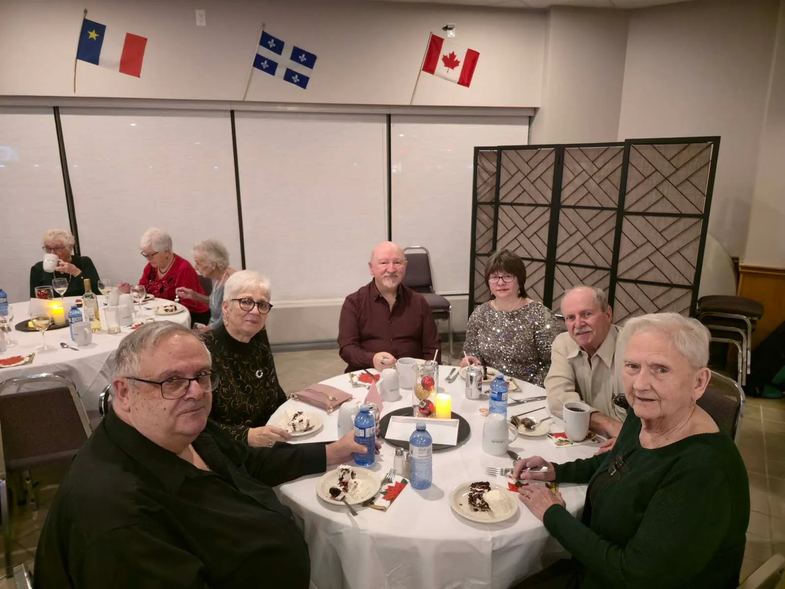 Groupe de membres assis à une table avec des assiettes de gâteau, sous les drapeaux du Québec, du Canada et de l’Acadie accrochés au mur.