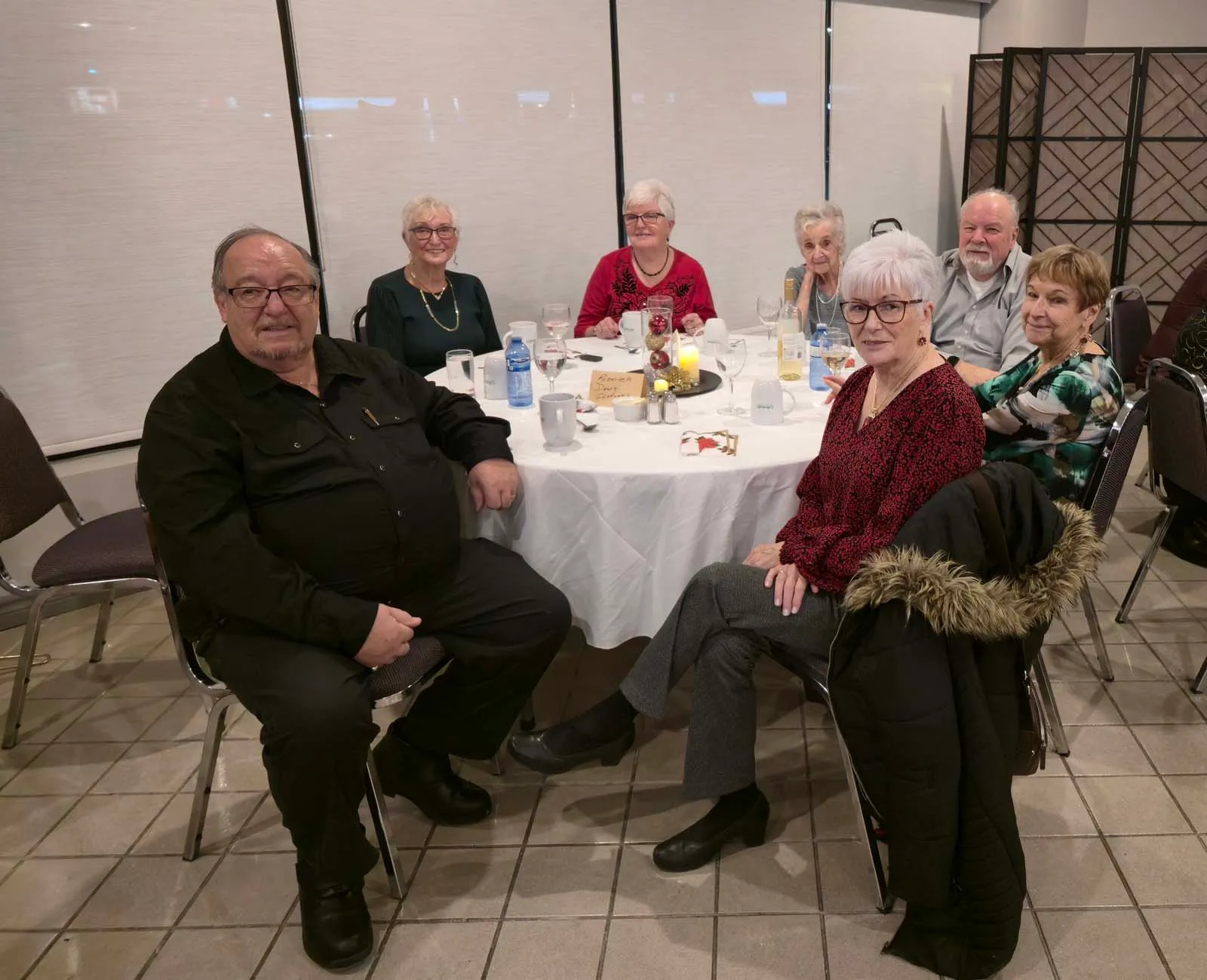 Sept membres de L’Amicale assis autour d’une table ronde, souriants après le repas, avec des tasses, verres et décorations au centre.