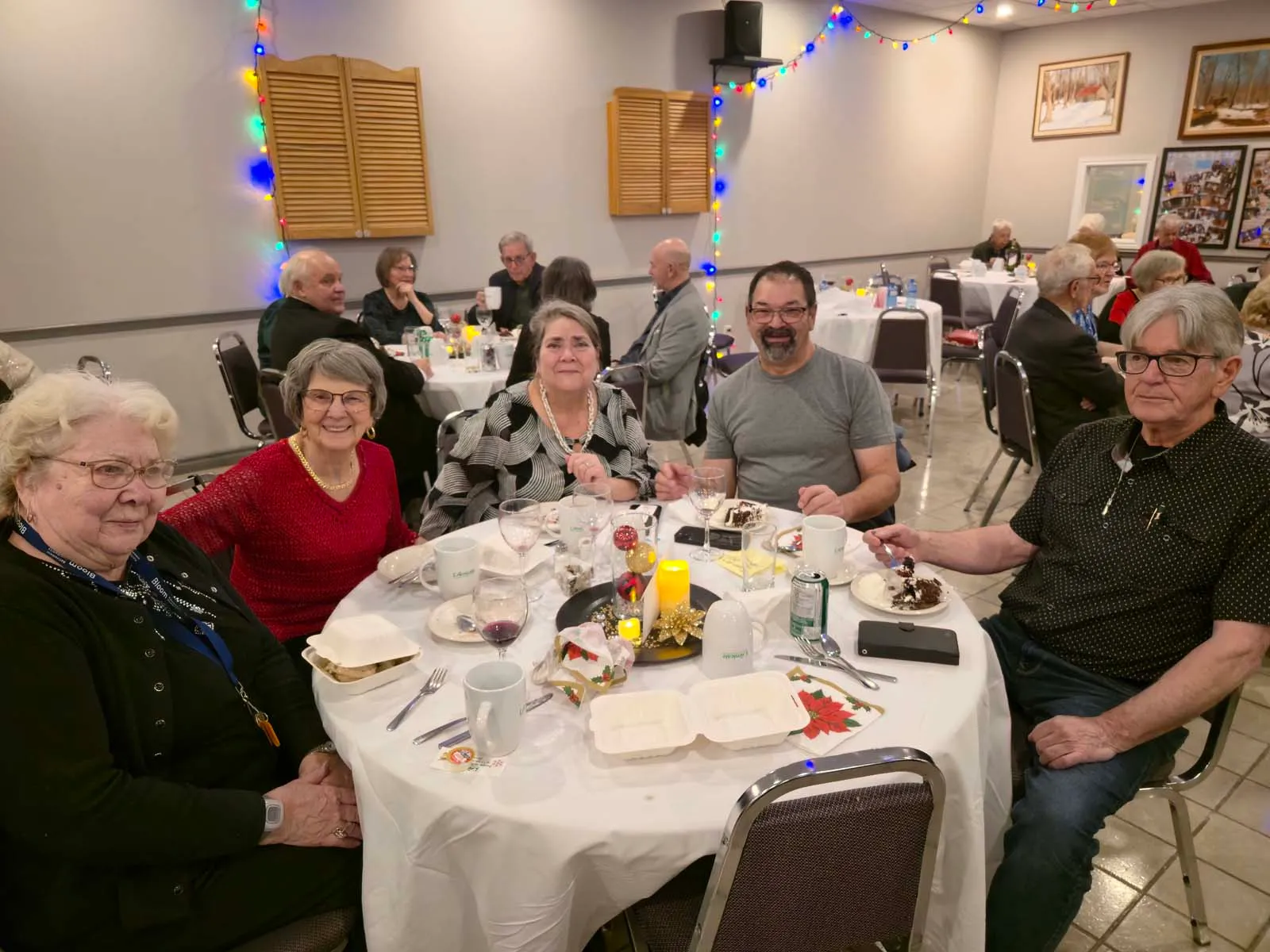 Groupe de membres de L’Amicale assis autour d’une table ronde, souriant à la caméra lors d’un repas festif dans une salle décorée de lumières.