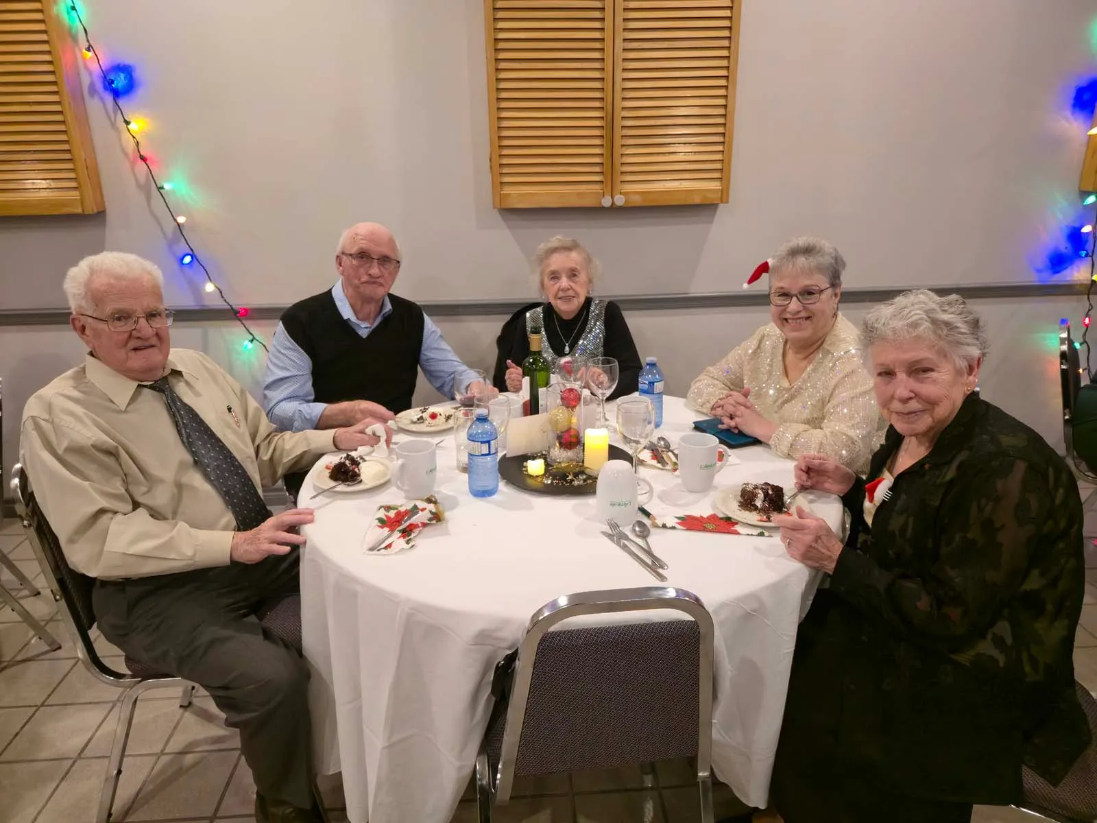 Cinq membres âgés de L’Amicale dégustant un dessert à une table élégamment dressée, entourée de guirlandes lumineuses multicolores.