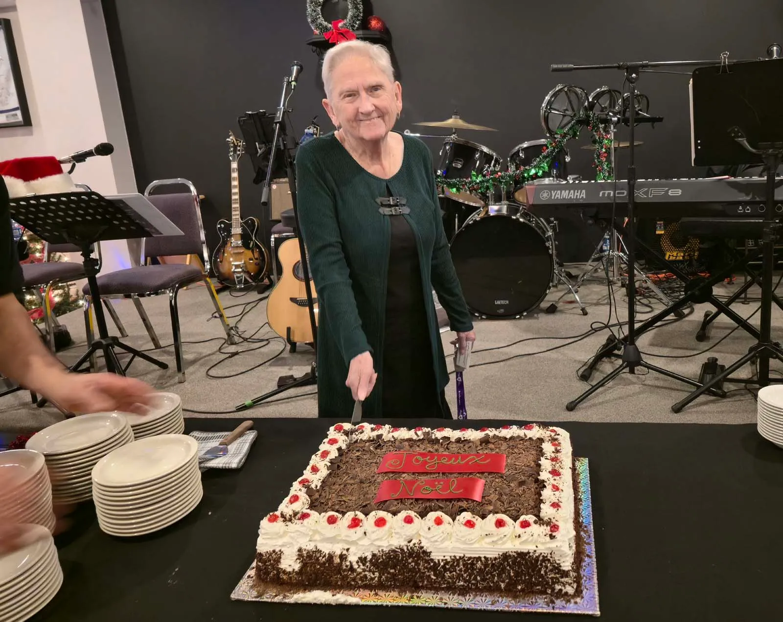 Une membre de L’Amicale souriante en train de couper le gâteau de Noël, devant une scène décorée avec des instruments de musique.