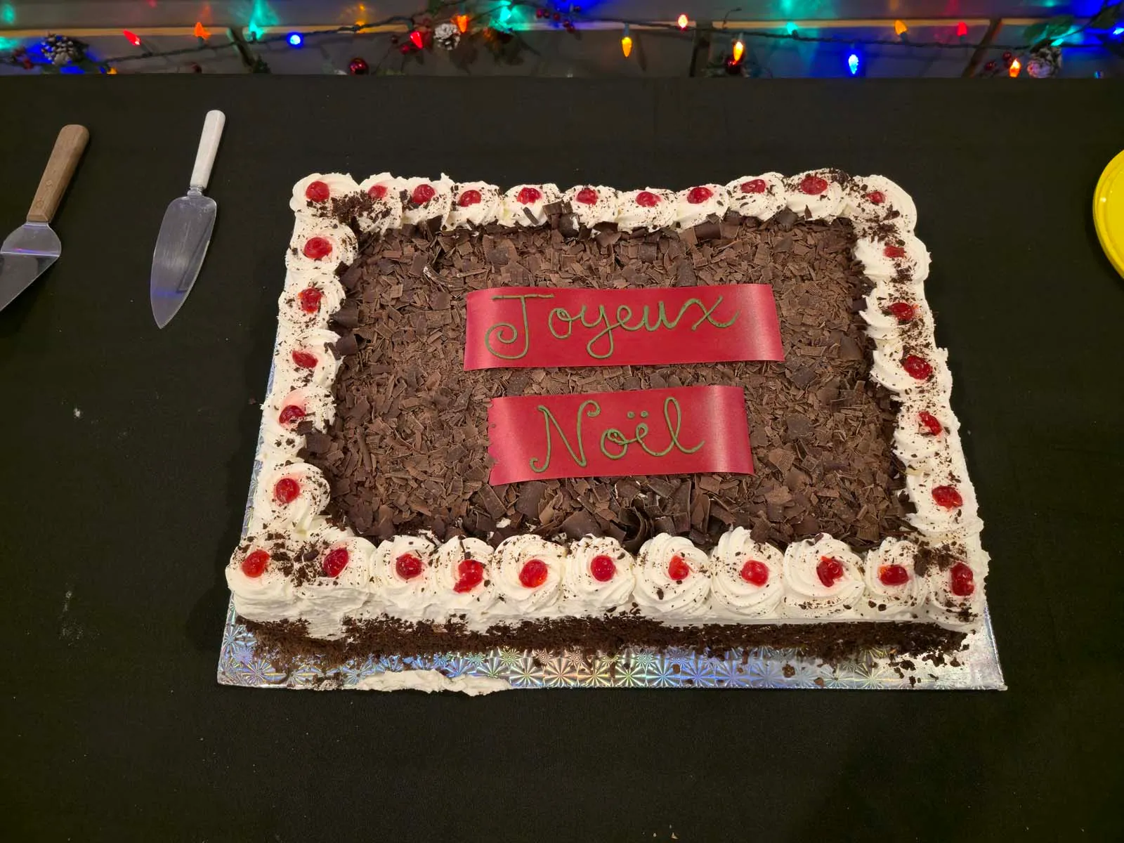 Gâteau rectangulaire au chocolat décoré de crème fouettée et de cerises, avec les mots "Joyeux Noël" écrits sur deux bandeaux rouges au centre.
