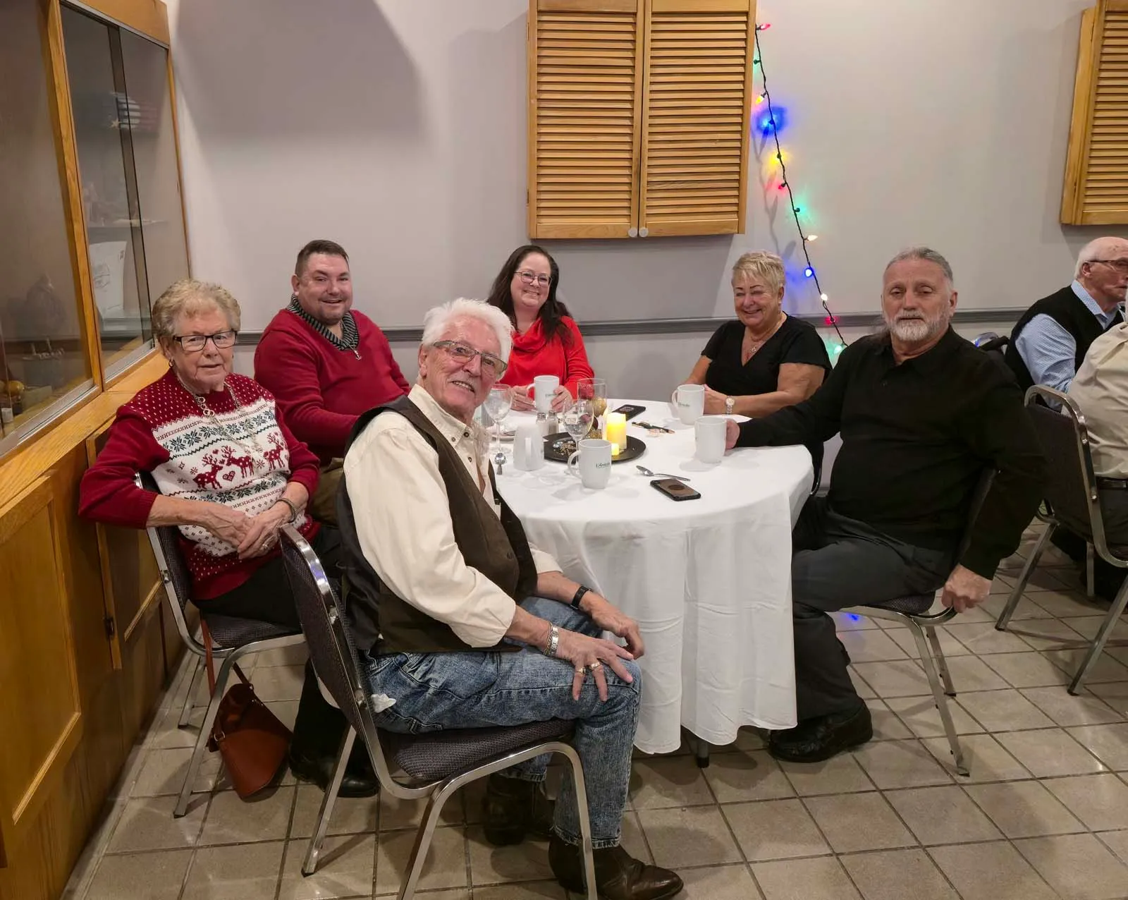 Groupe de six membres de L’Amicale réunis autour d’une table décorée, avec des guirlandes lumineuses accrochées au mur en arrière-plan.