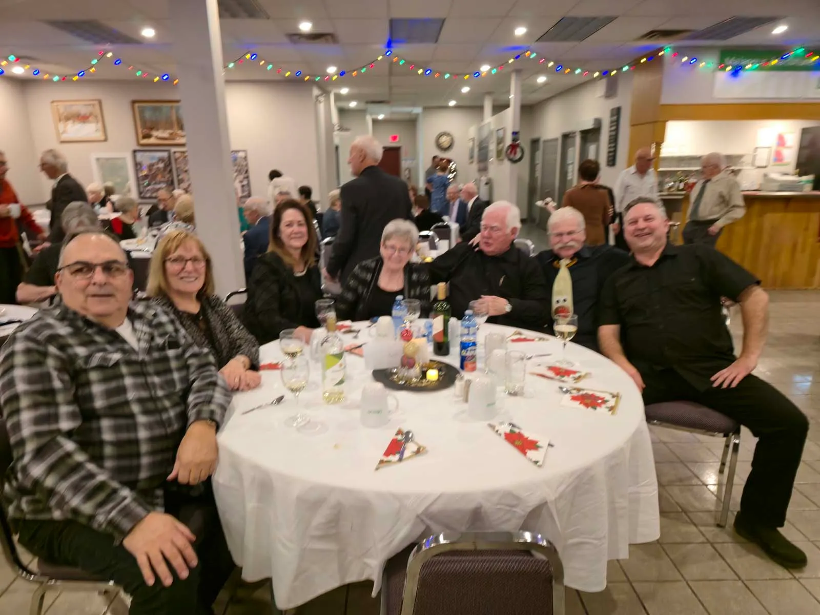 Table de huit personnes en ambiance festive, certains membres regardent l’objectif tandis que d’autres discutent entre eux, avec des décorations suspendues.