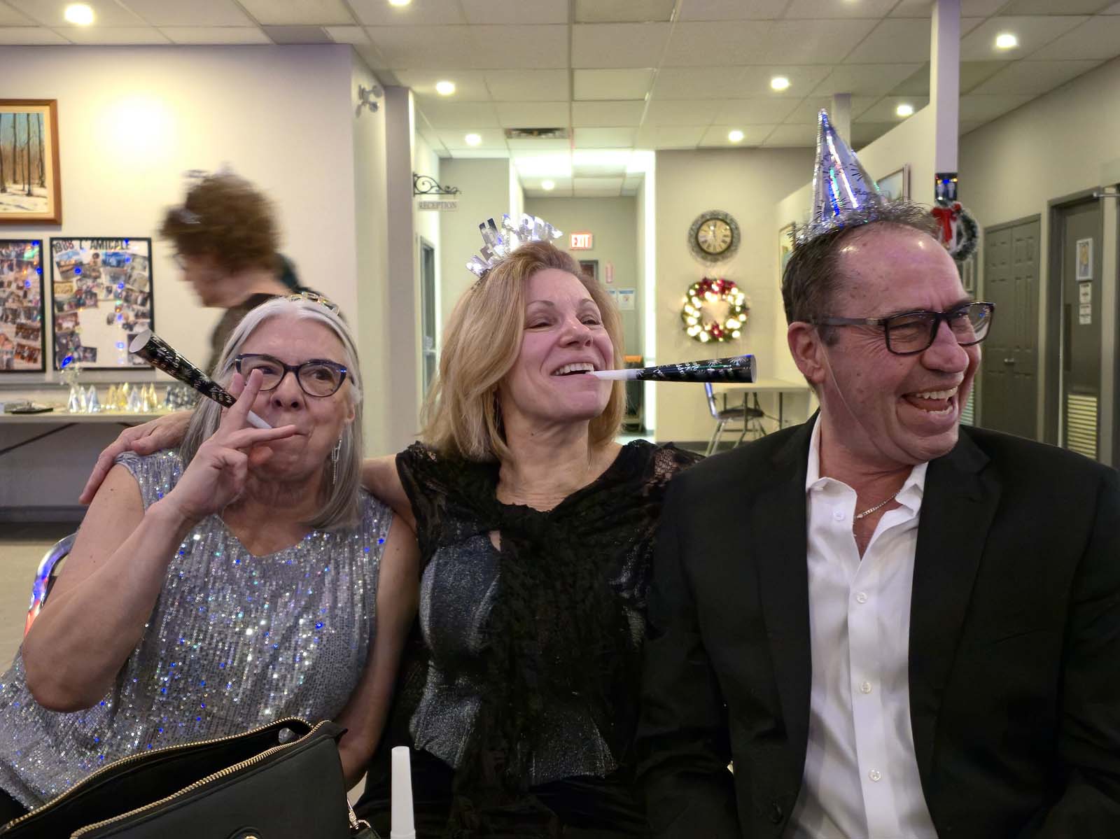 Trois membres de L’Amicale assis ensemble, riant et célébrant le Nouvel An avec des accessoires festifs dans une ambiance joyeuse.