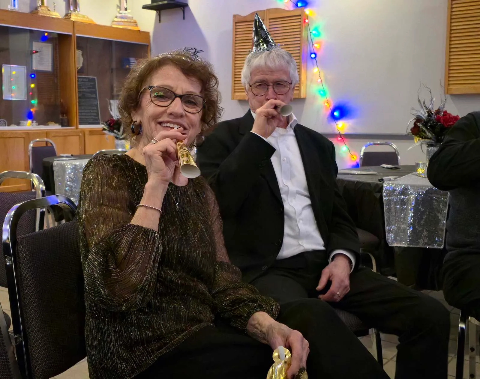 Deux membres de L’Amicale célébrant le Nouvel An, soufflant dans des sans-gênes et souriant à la caméra.