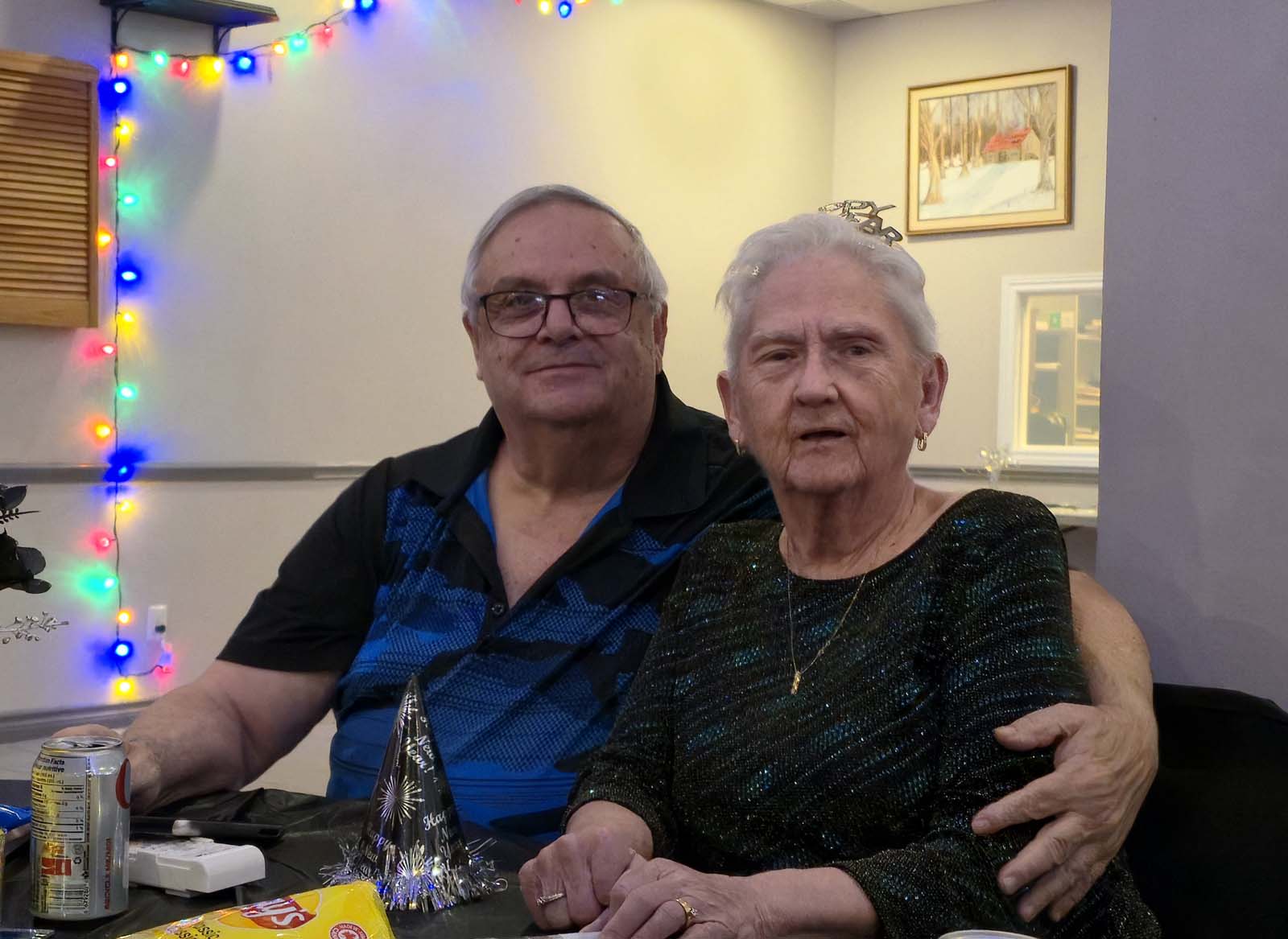 Deux membres de L’Amicale assis côte à côte, souriant à la caméra lors de la soirée du Nouvel An, avec décorations lumineuses en arrière-plan