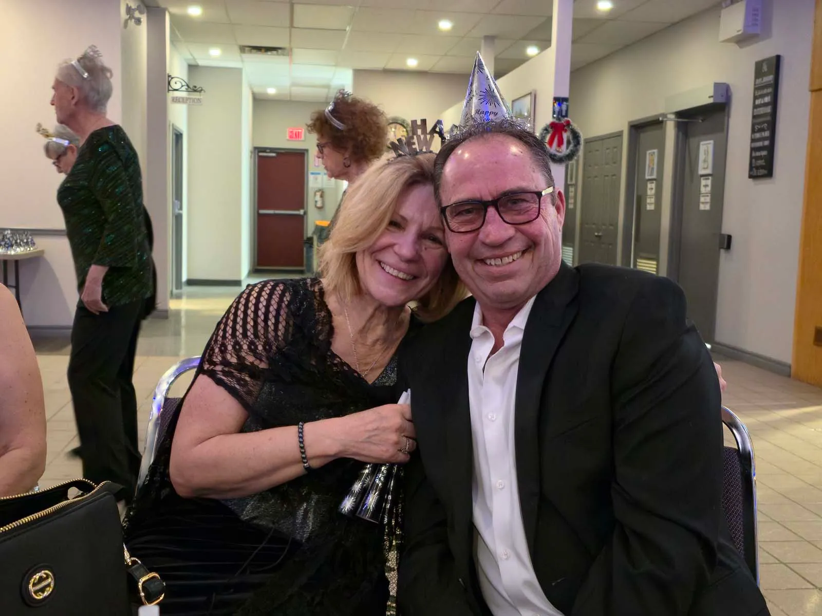 Deux membres de L’Amicale assis côte à côte, souriant à la caméra, avec des décorations festives visibles derrière eux.