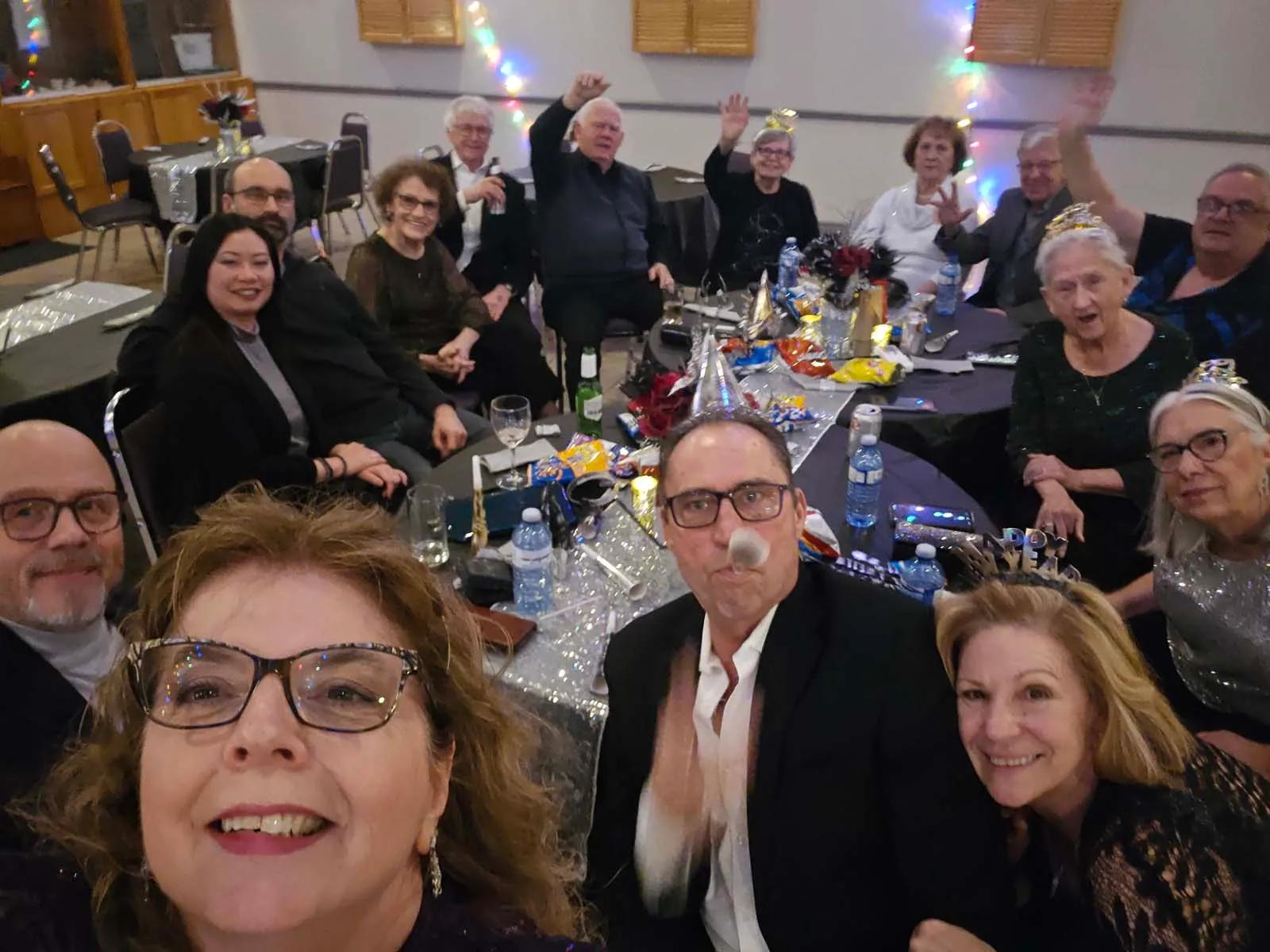Groupe de membres de L’Amicale rassemblés autour d’une table, souriant et saluant la caméra lors de la célébration du Nouvel An.