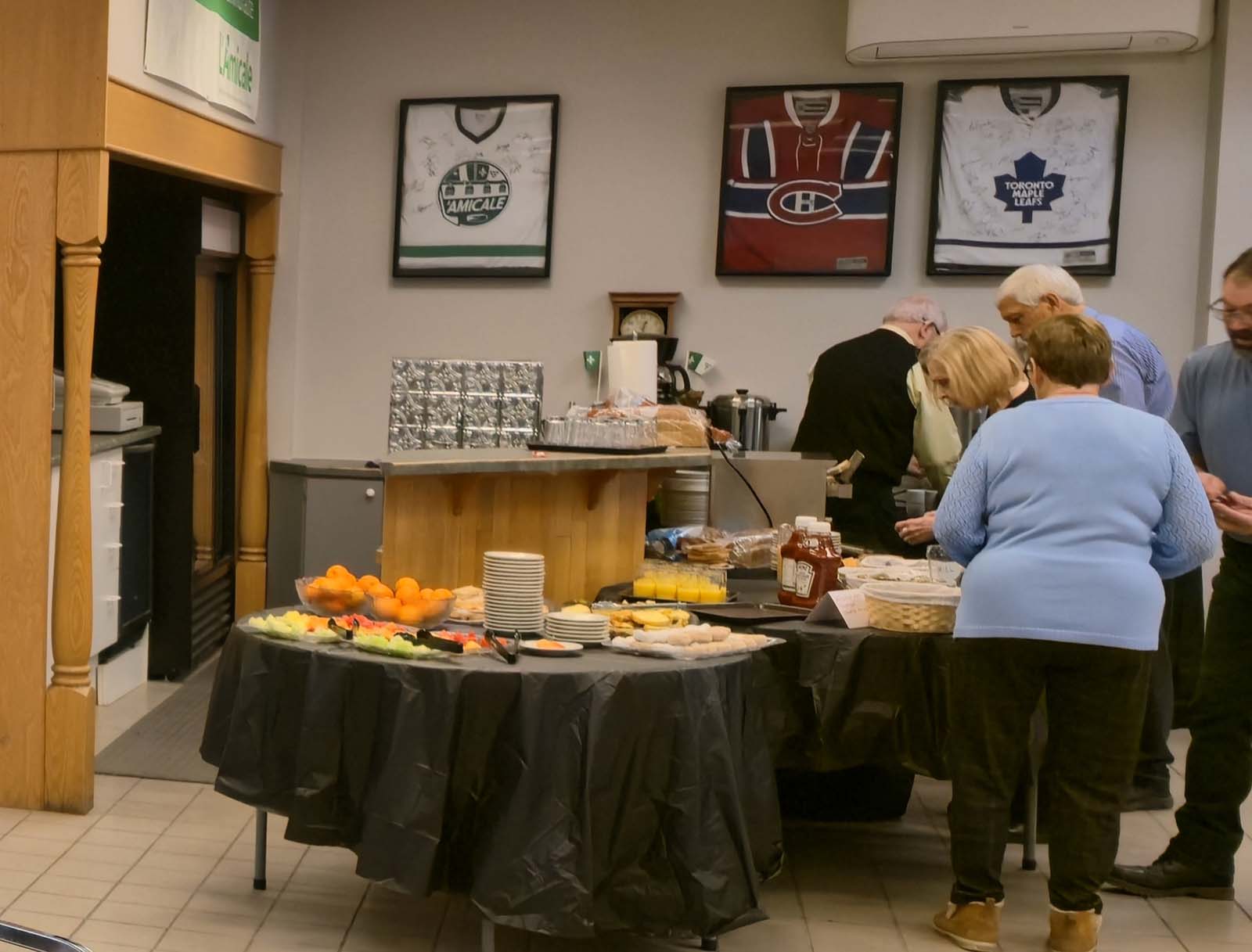 Table de service du repas à L’Amicale, avec des membres se servant nourriture et boissons sous des chandails sportifs encadrés au mur.